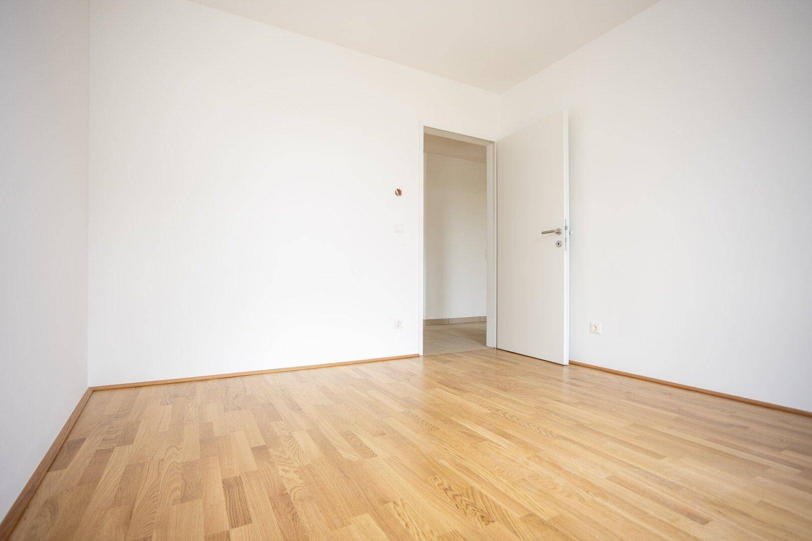 Geräumiges Zimmer mit hellem Parkettboden und weiß gestrichenen Wänden, mit Türzugang.