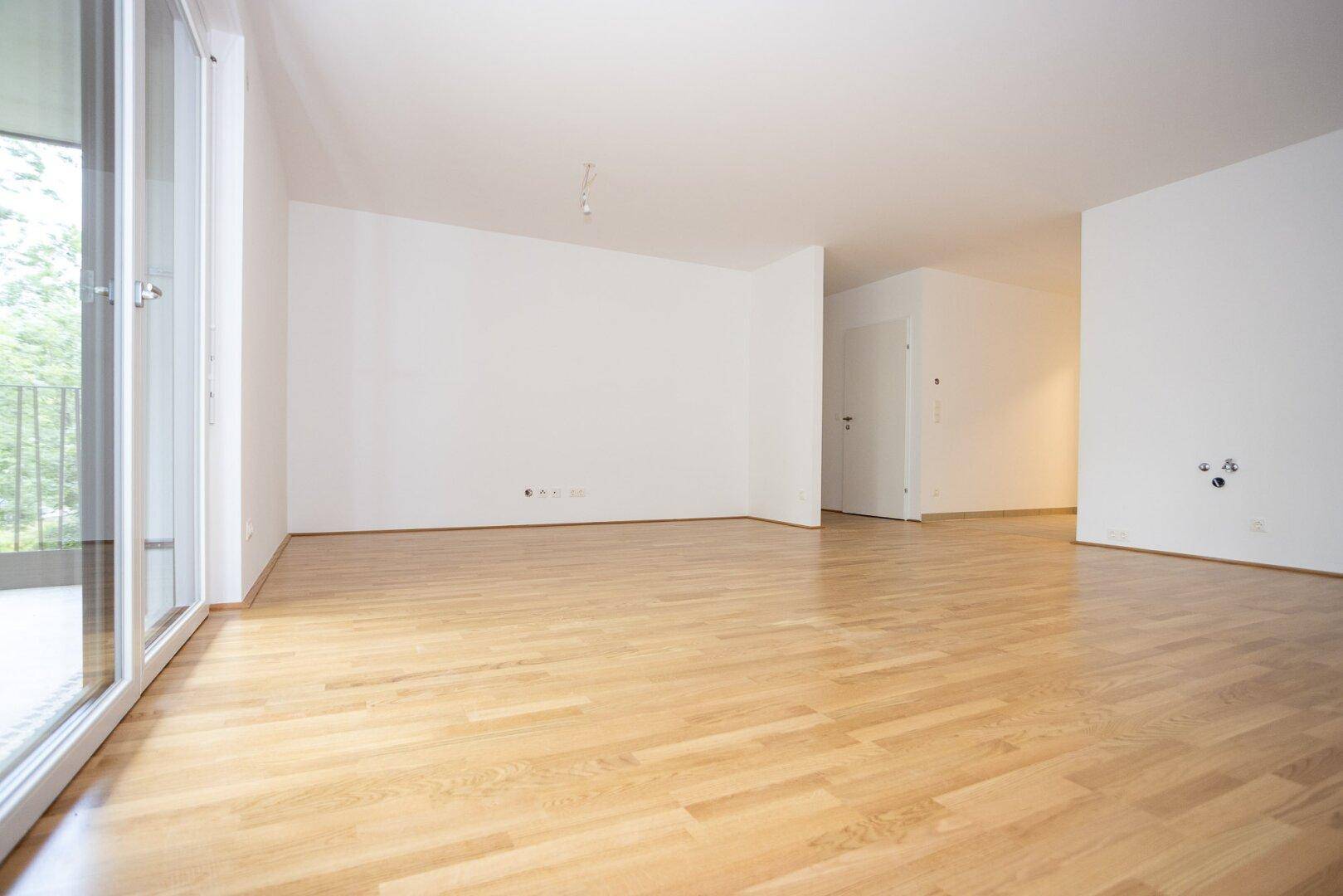 Geräumiges, unmöbliertes Zimmer mit hellem Holzfußboden und viel Tageslicht durch die Balkontür.