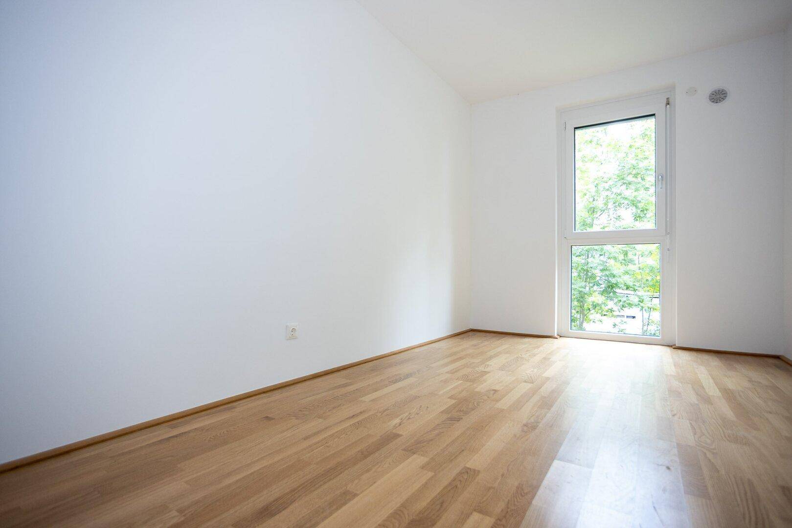 Ruhiges Schlafzimmer mit hellem Parkettboden und einem großen Fenster, das viel Licht hereinlässt.