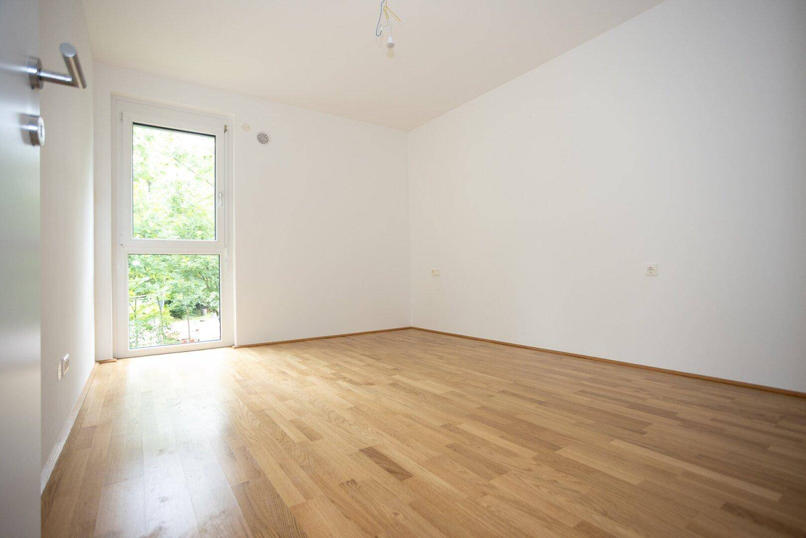 Unmöbliertes Zimmer mit glänzendem Parkettboden und einem Fenster mit Blick ins Grüne.