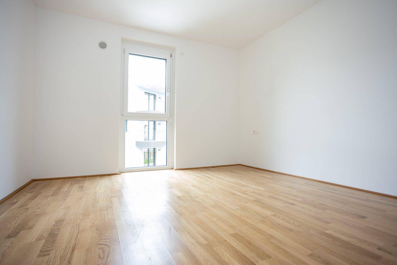Freundliches Zimmer mit Holzboden und großem Fenster, das viel Tageslicht hereinlässt.