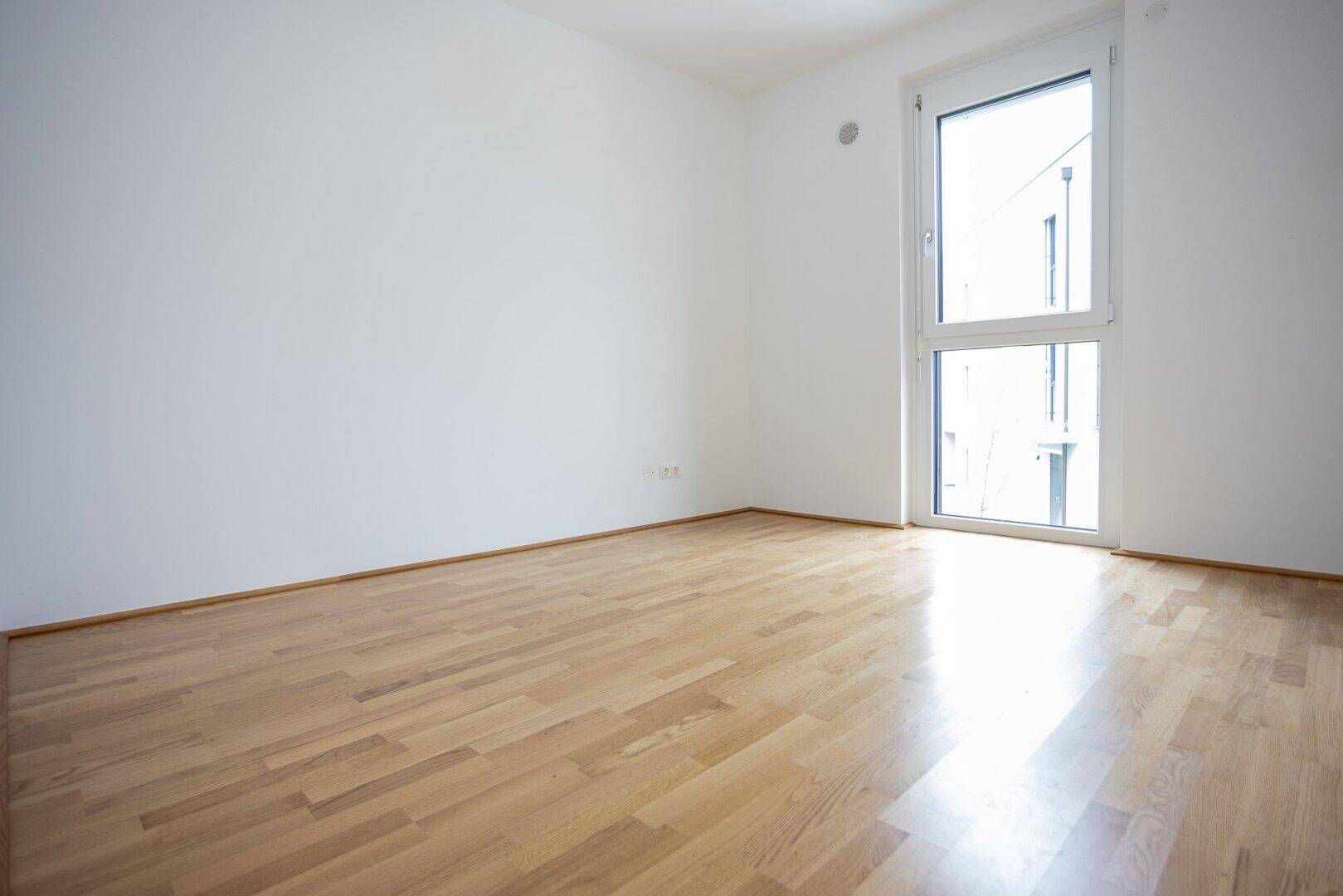 Heller Raum mit Parkettboden und einem hohen Fenster, das eine angenehme Atmosphäre schafft.