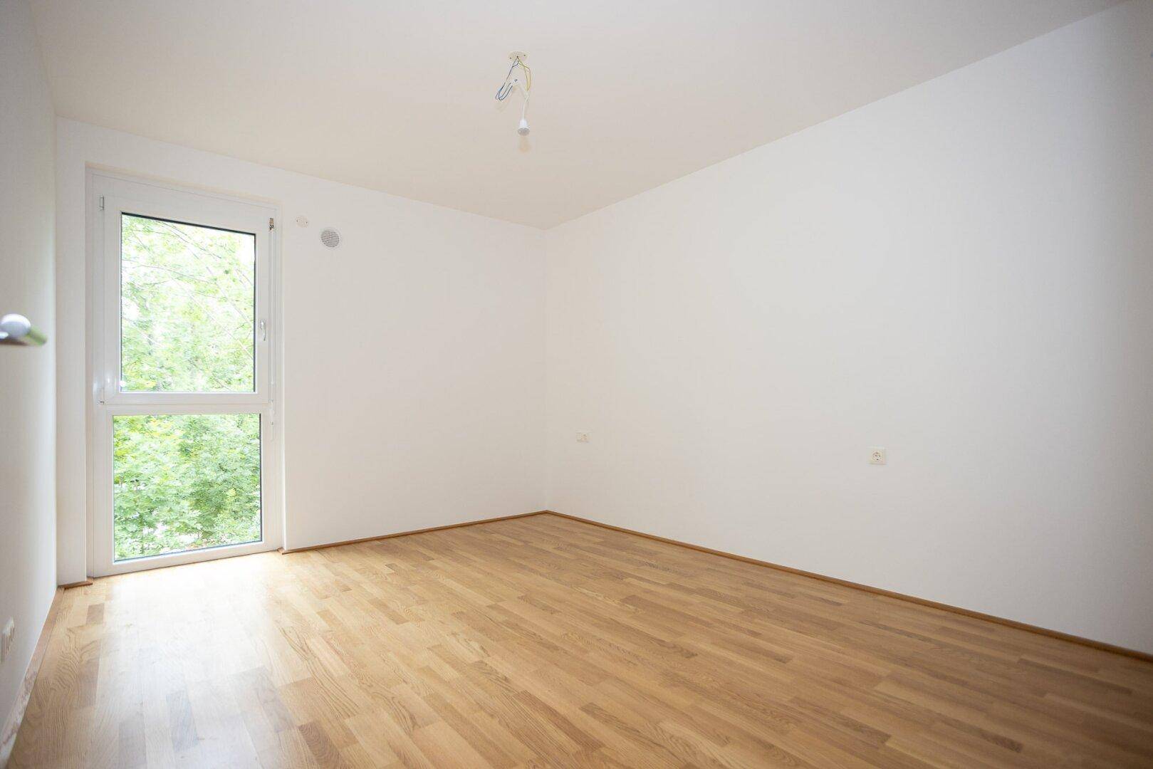 Schlafzimmer mit Parkettboden und großem Fenster, das viel Tageslicht und einen grünen Ausblick bietet.