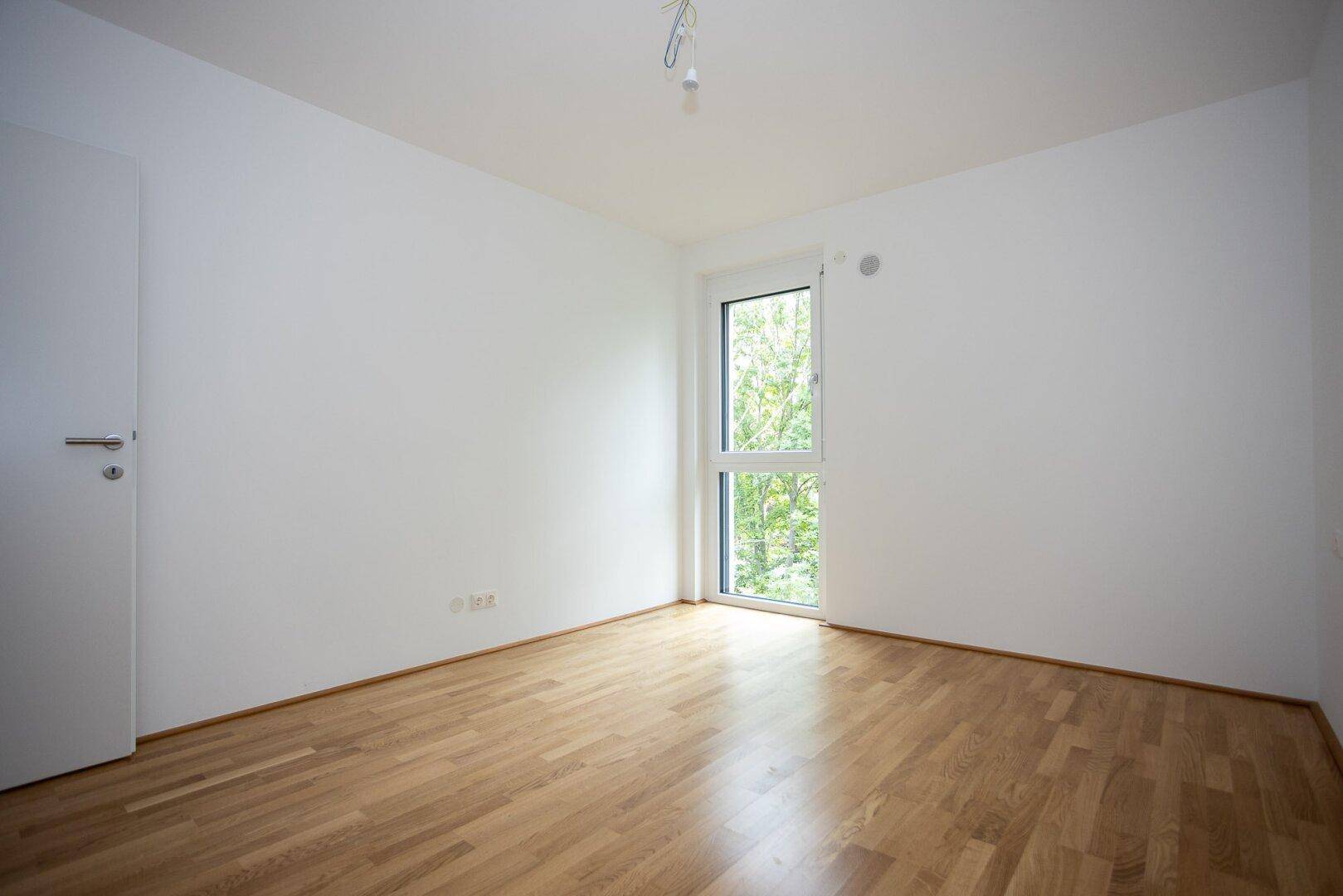Helles Zimmer mit Parkettboden und Fenster mit Blick auf Bäume, ideal als Schlaf- oder Arbeitszimmer.