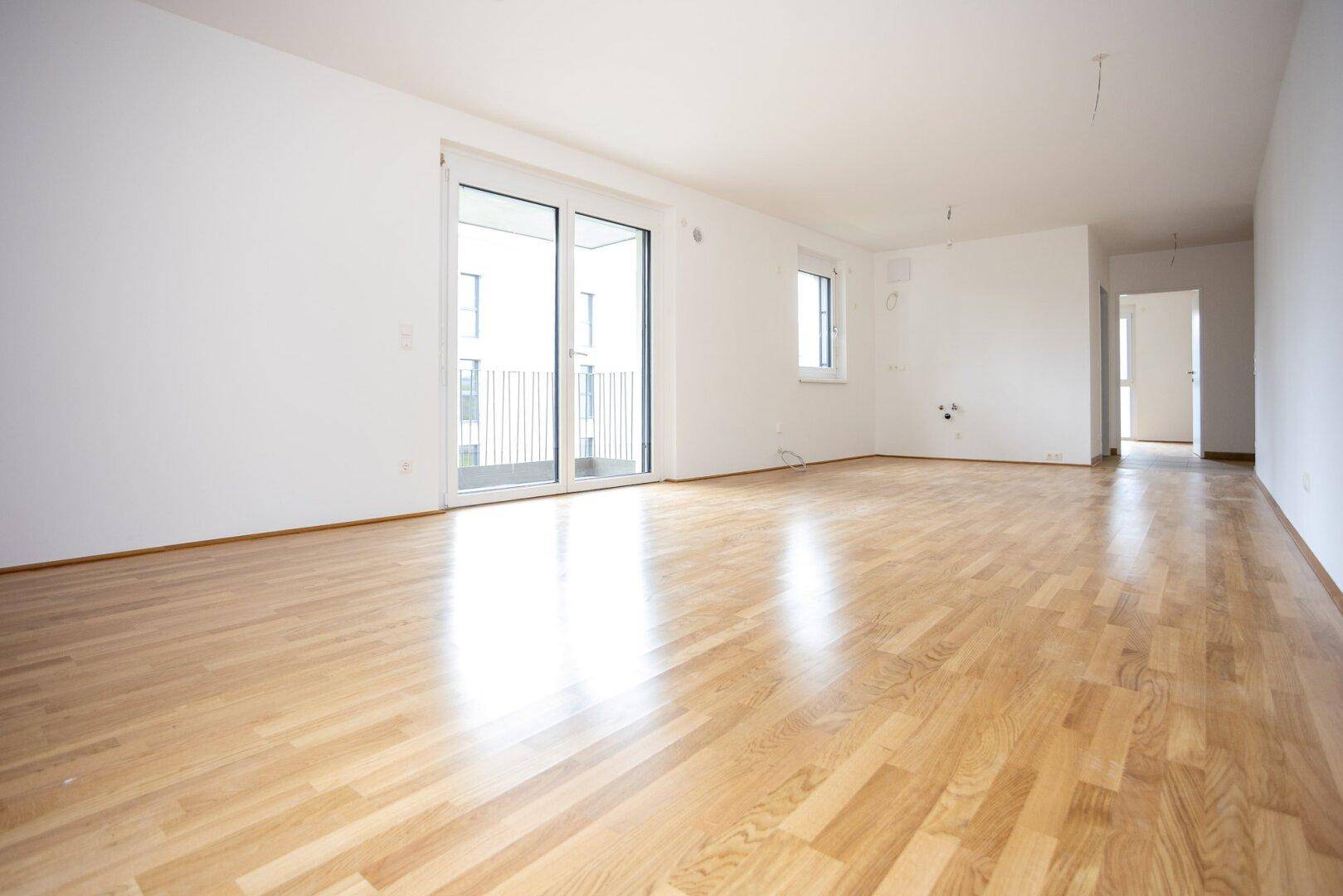 Geräumiges, unmöbliertes Zimmer mit hellem Holzfußboden, großen Fenstern und Zugang zum Balkon.