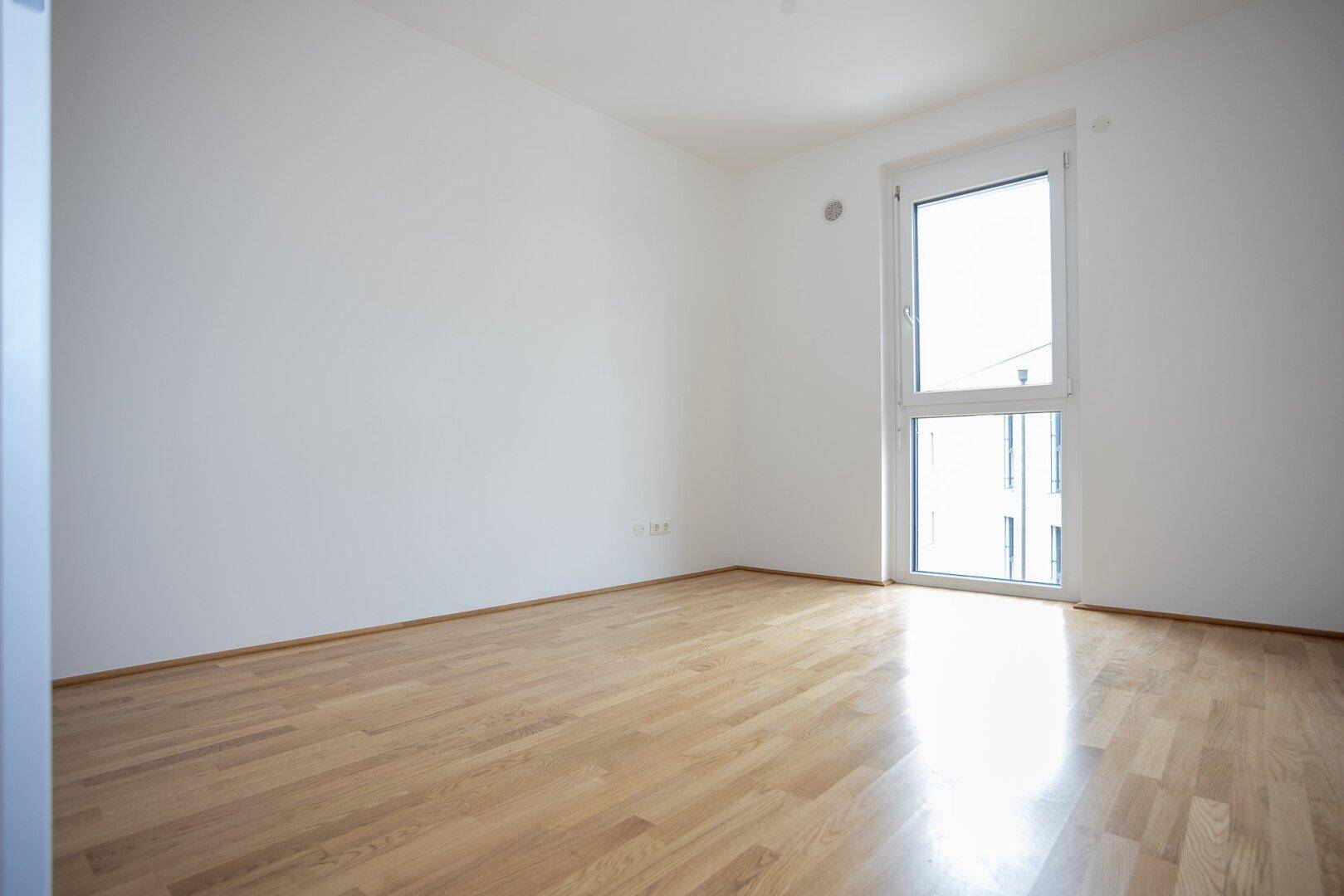 Helles Schlafzimmer mit Holzparkett und einem hohen Fenster, das viel Tageslicht hereinlässt.