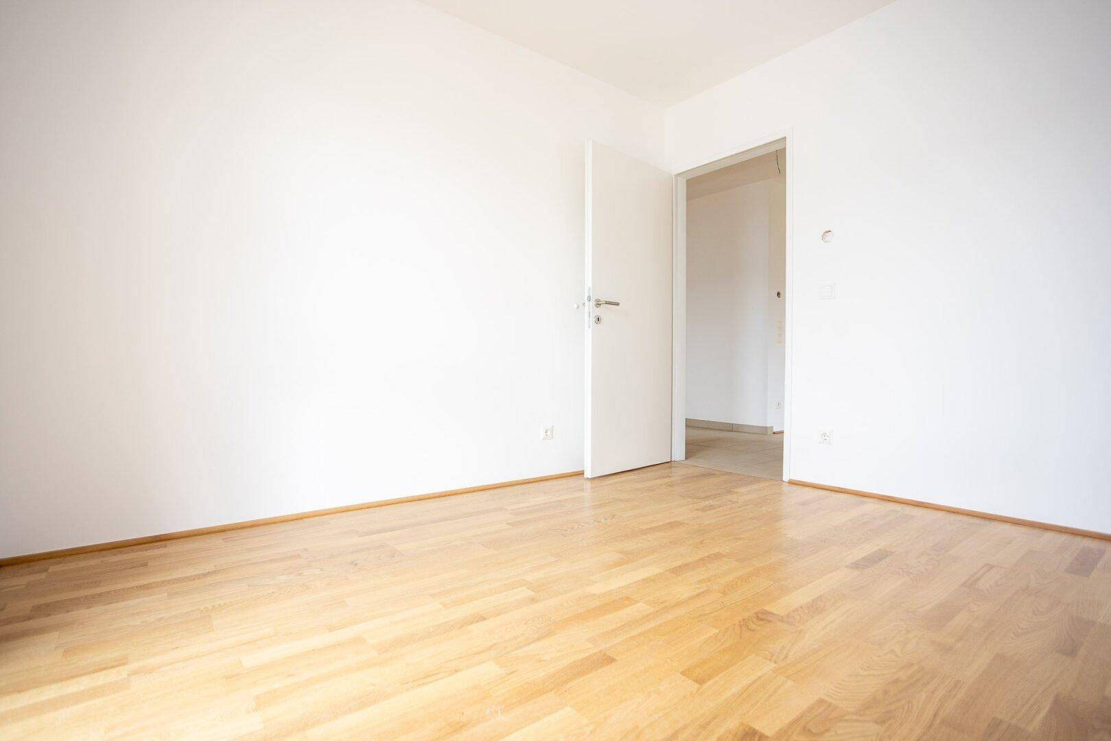 Leeres Zimmer mit hellem Holzfußboden und weiß gestrichenen Wänden, bietet Zugang zu einem weiteren Raum.