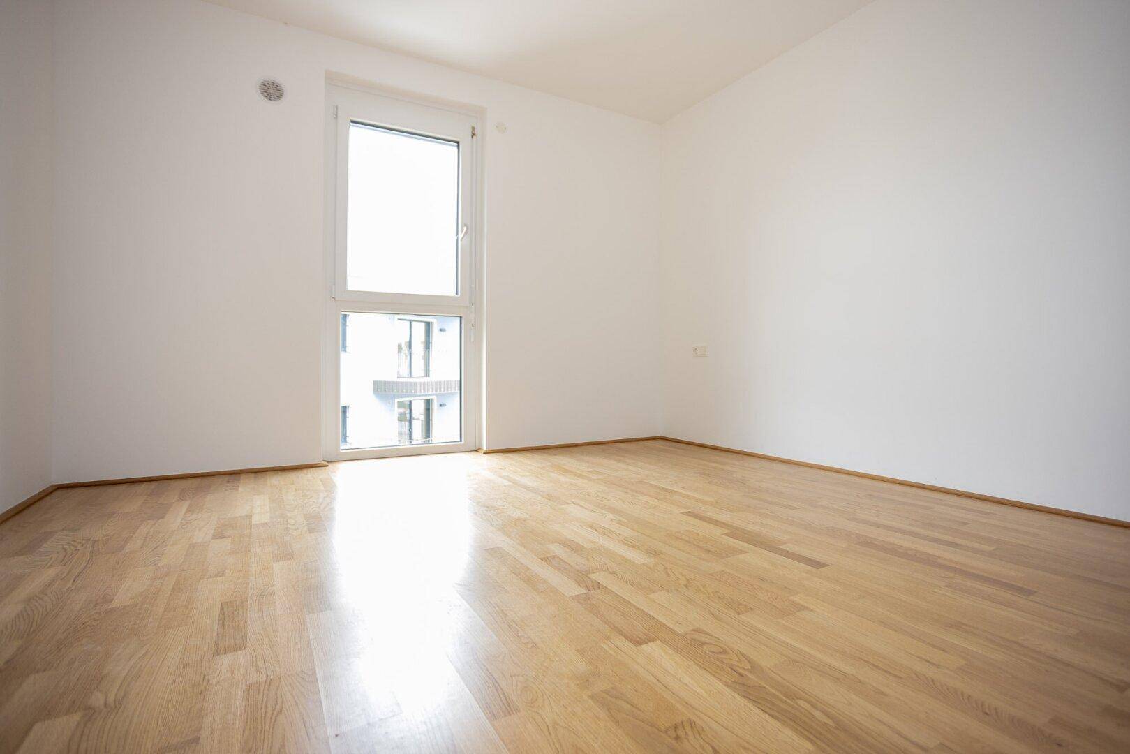 Unmöbliertes Schlafzimmer mit glänzendem Holzparkett und einem hohen Fenster, das den Raum erhellt.