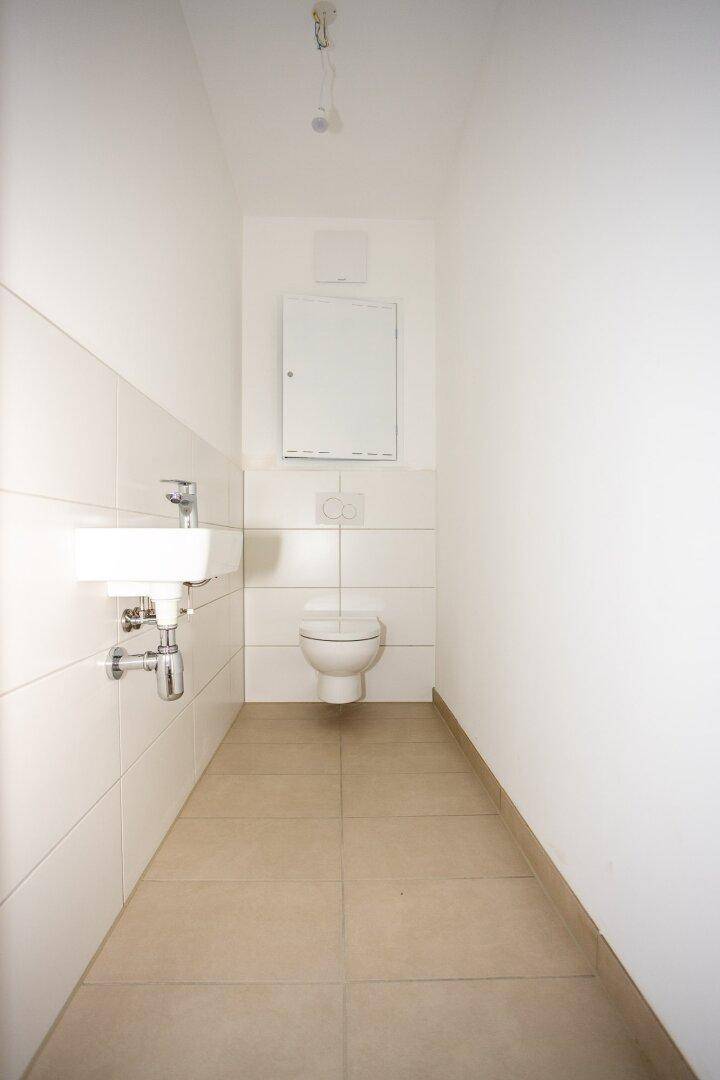 Schmale Gästetoilette mit Wand-WC, kleinem Waschbecken und Spiegelschrank, hell gefliest.