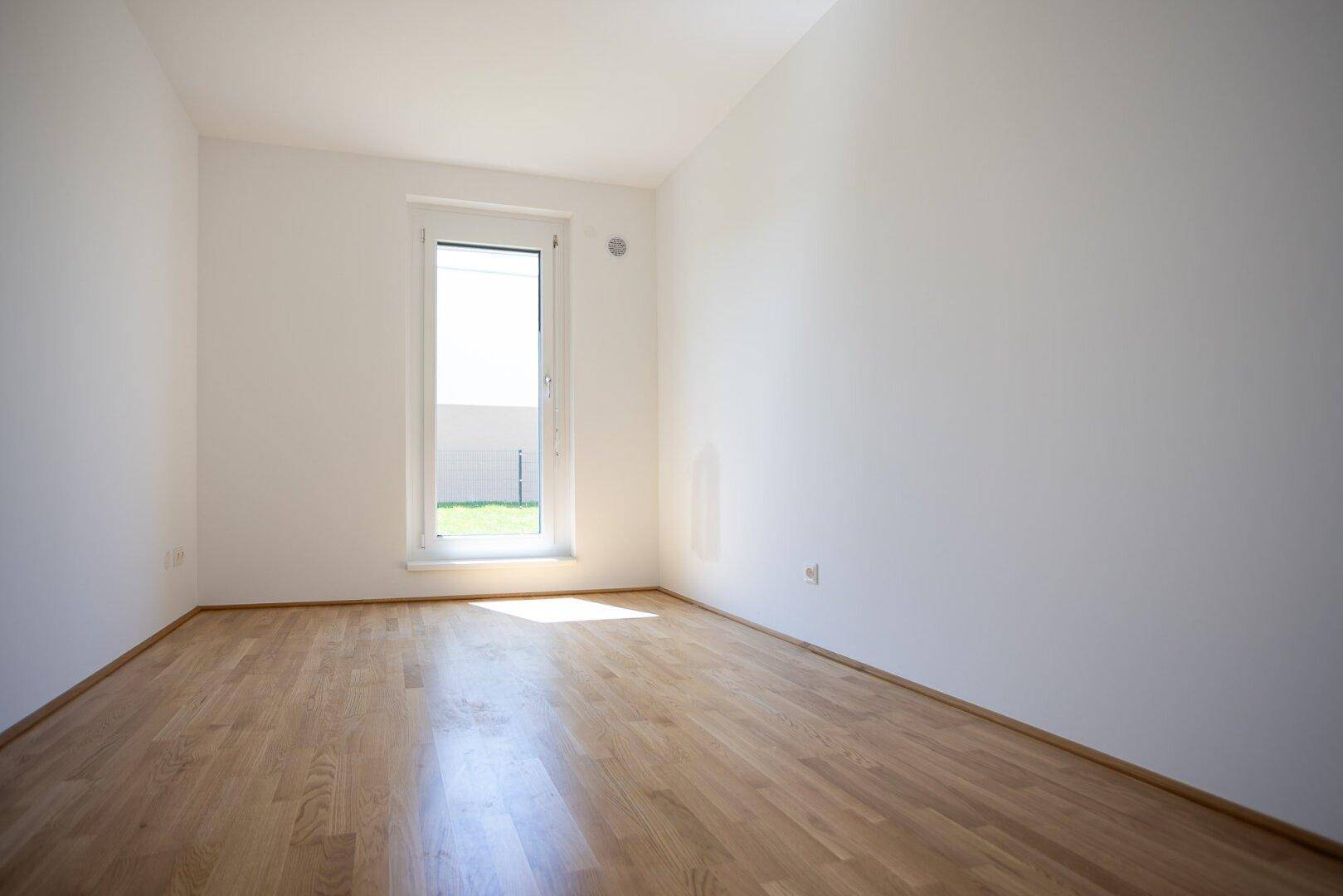 Sonniger Raum mit Holzboden und einem hohen Fenster, das den Blick ins Grüne freigibt.