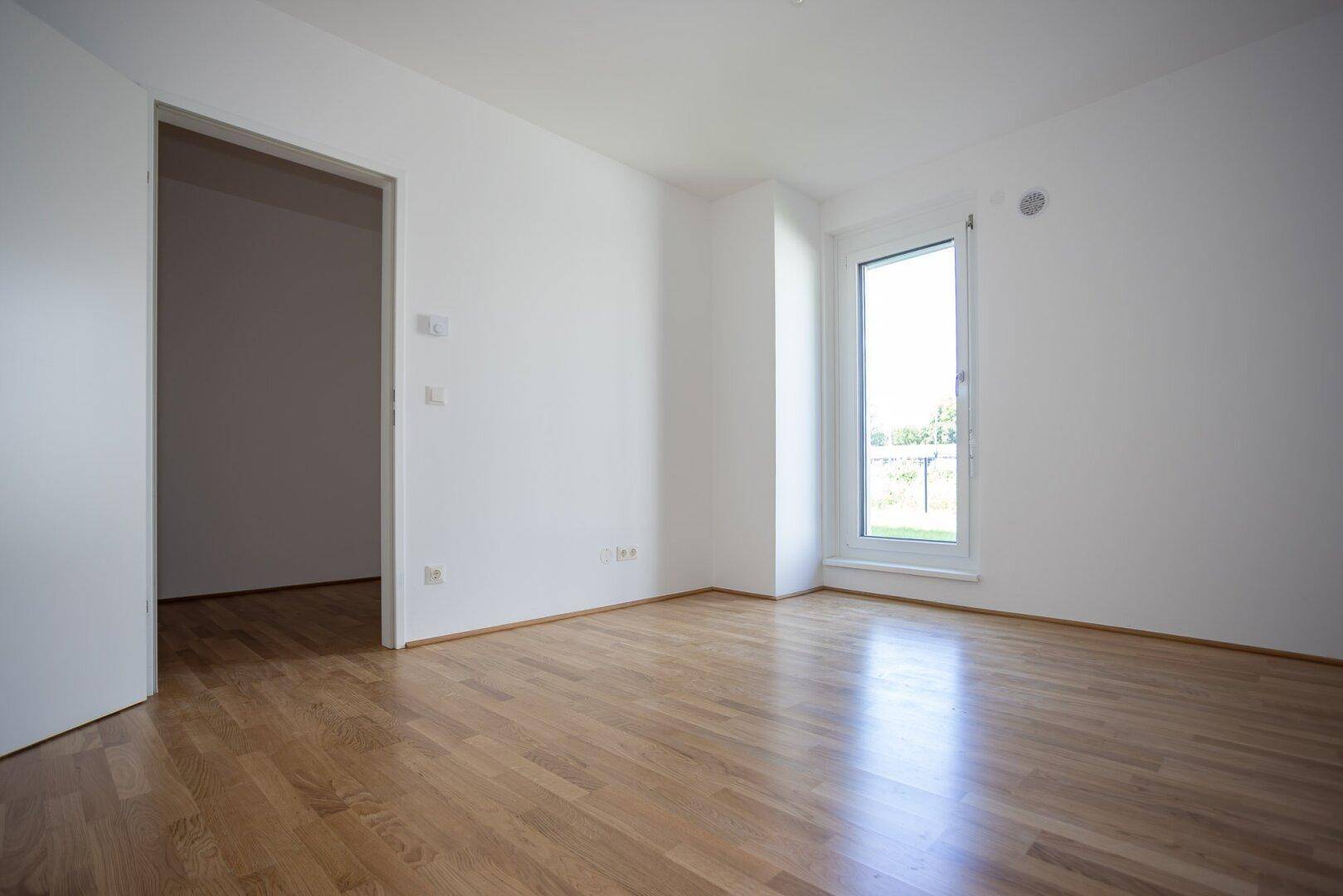 Geräumiges, helles Zimmer mit Holzboden und weiß gestrichenen Wänden, mit Türzugang.