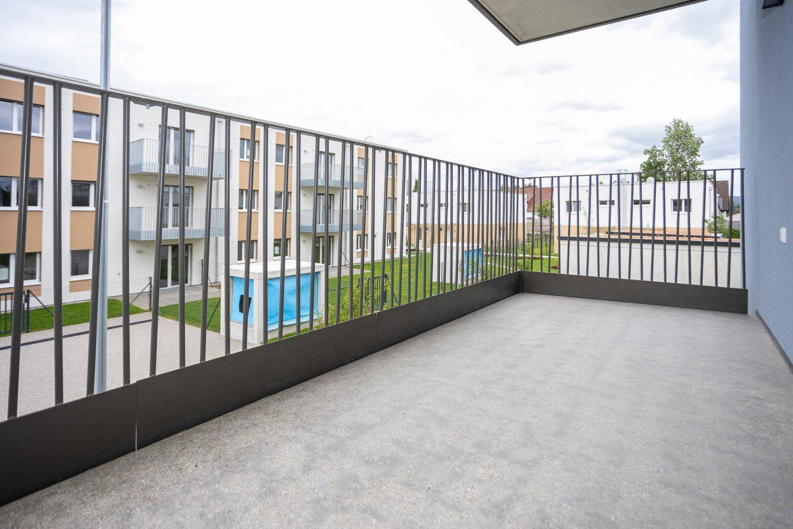 Geräumiger Balkon mit Betonboden und Metallgeländer, bietet Blick auf die Wohnanlage.