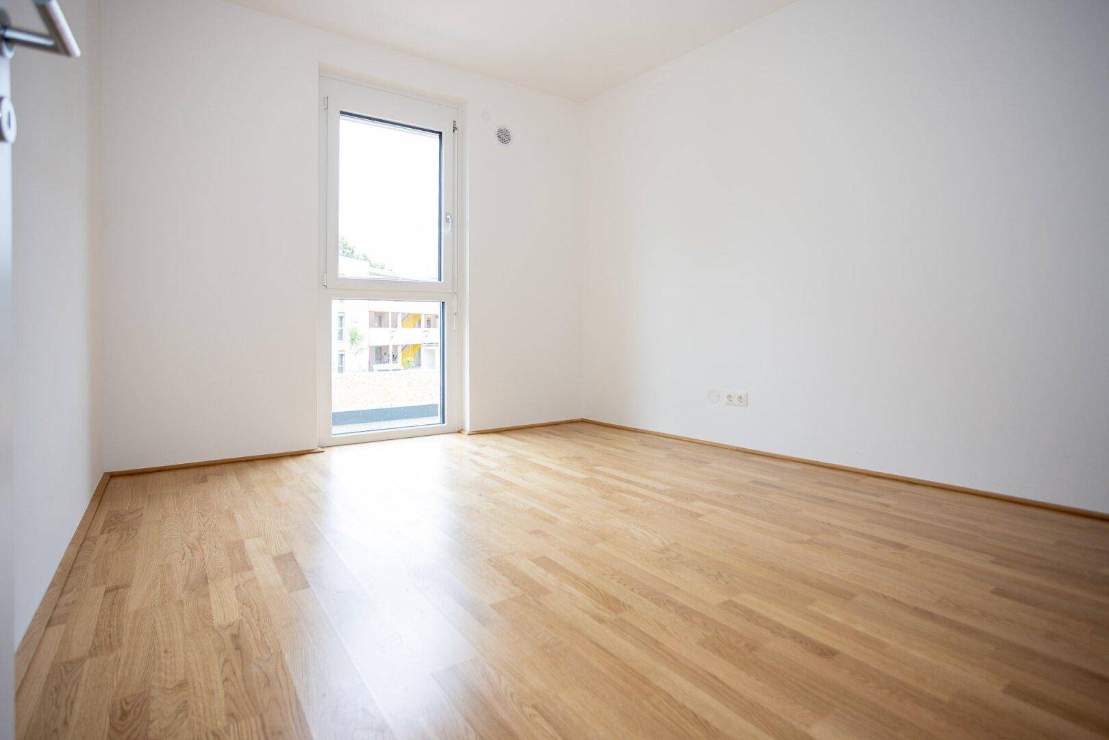 Helles Schlafzimmer mit Parkettboden und großem Fenster, ideal für natürliche Belichtung.