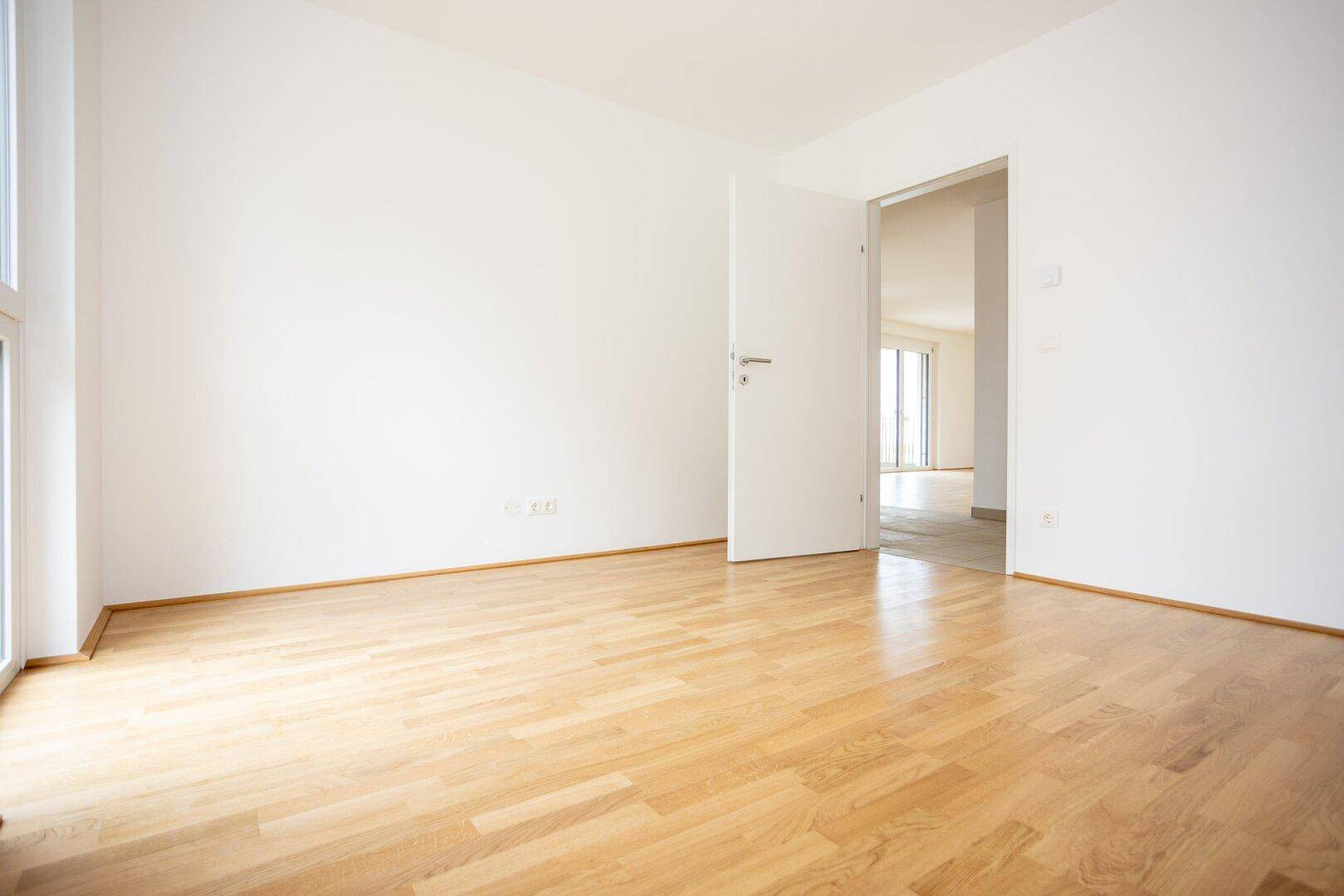 Leeres Schlafzimmer mit Parkettboden und Zugang zu einem weiteren Raum, ideal für Familien.