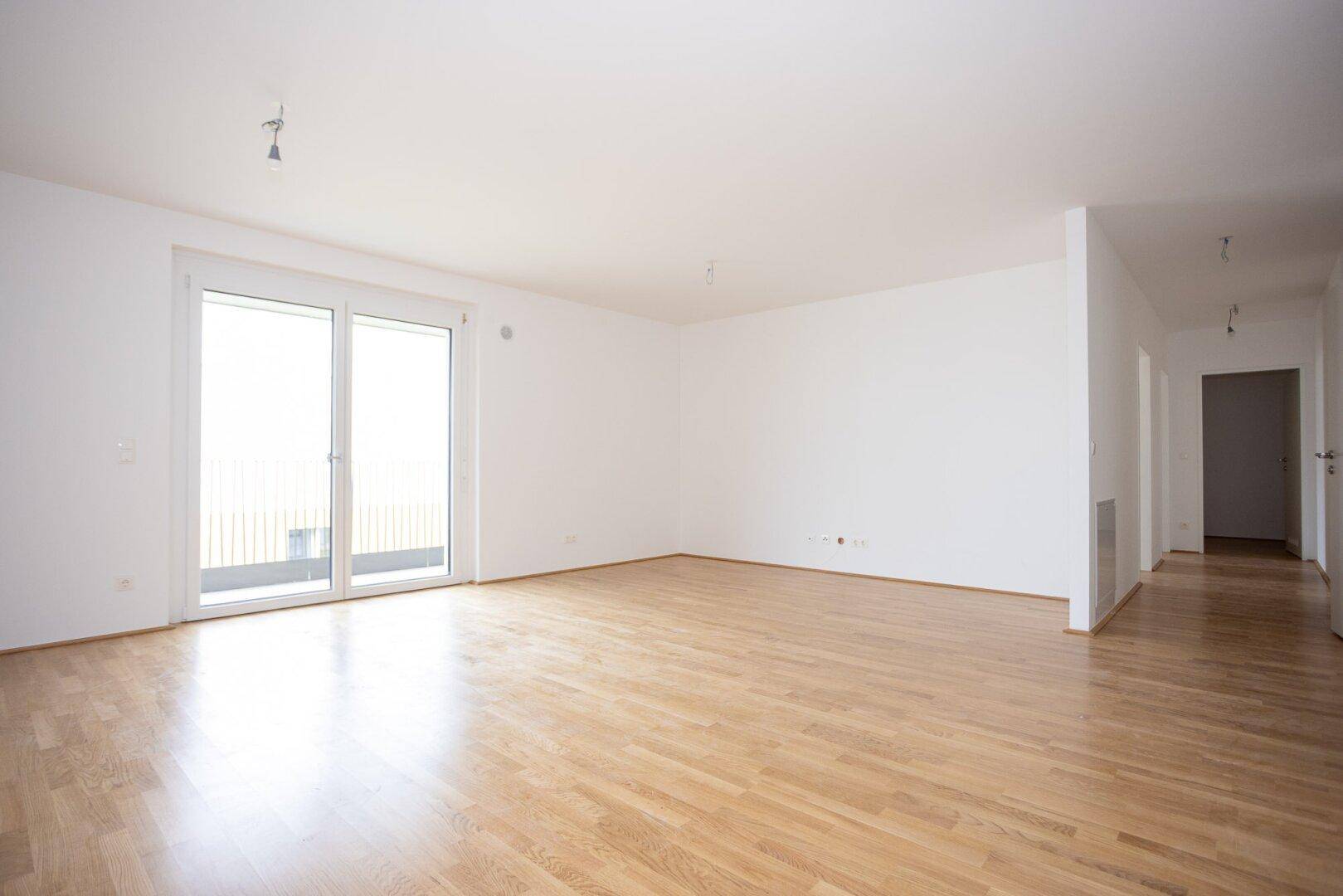 Geräumiges, leeres Wohnzimmer mit hellem Parkettboden und Balkonzugang, ideal für individuelle Gestaltung.