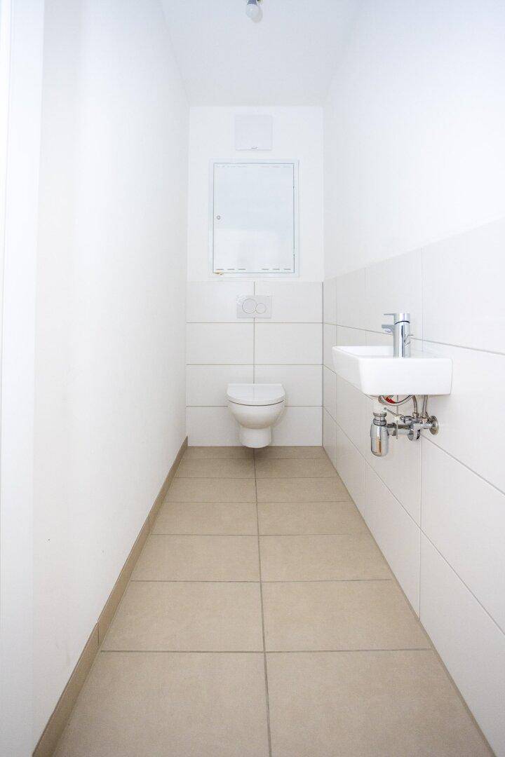 Separates WC mit wandhängender Toilette, kleinem Waschbecken und hellen Fliesen, funktional und sauber.
