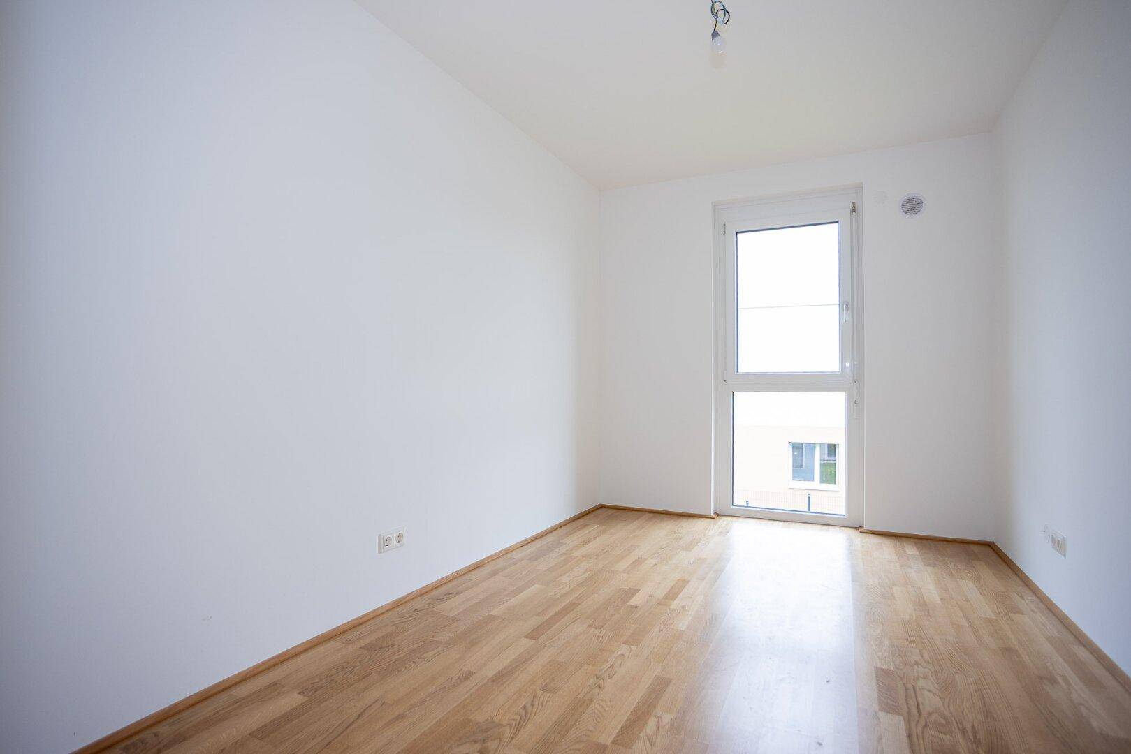 Helles Schlafzimmer mit Parkettboden und großem Fenster für viel Tageslicht, unmöbliert.