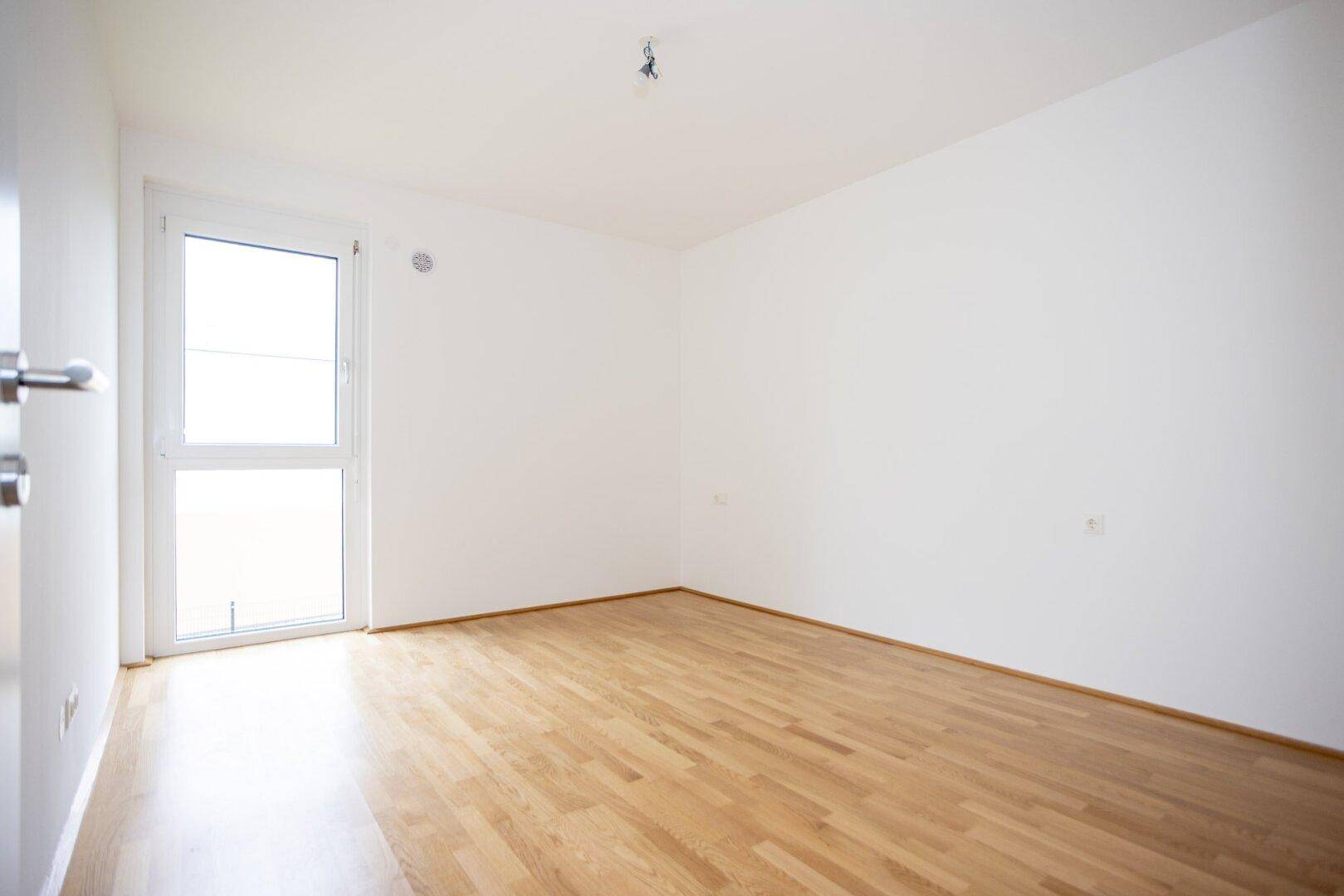Unmöbliertes Schlafzimmer mit hellem Parkettboden und Fenster mit Blick nach draußen, sehr einladend.