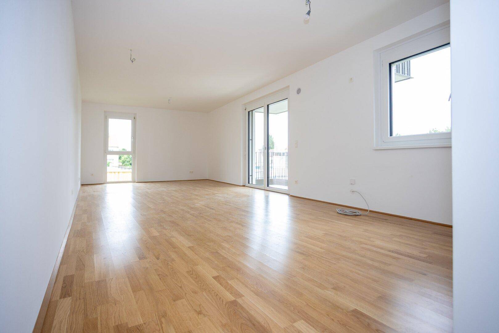 Geräumiges, helles Zimmer mit Holzboden und großen Fenstern, das viel Tageslicht bietet.