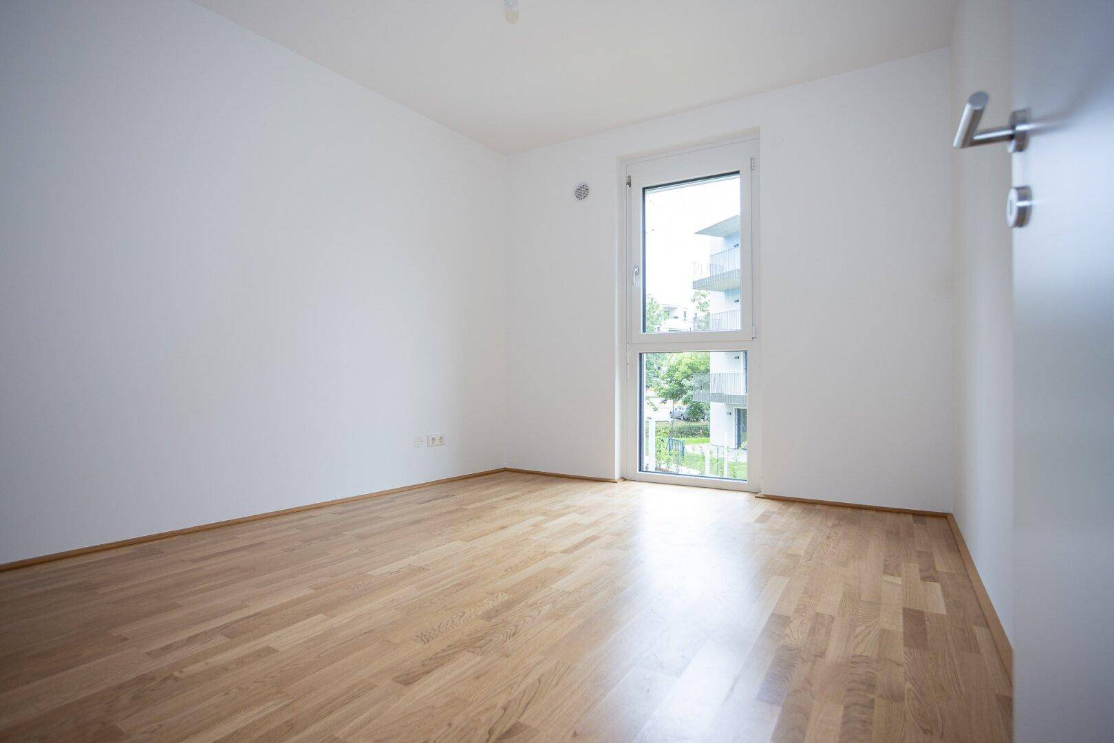 Helles Schlafzimmer mit Parkettboden und großem Fenster mit Blick ins Grüne.