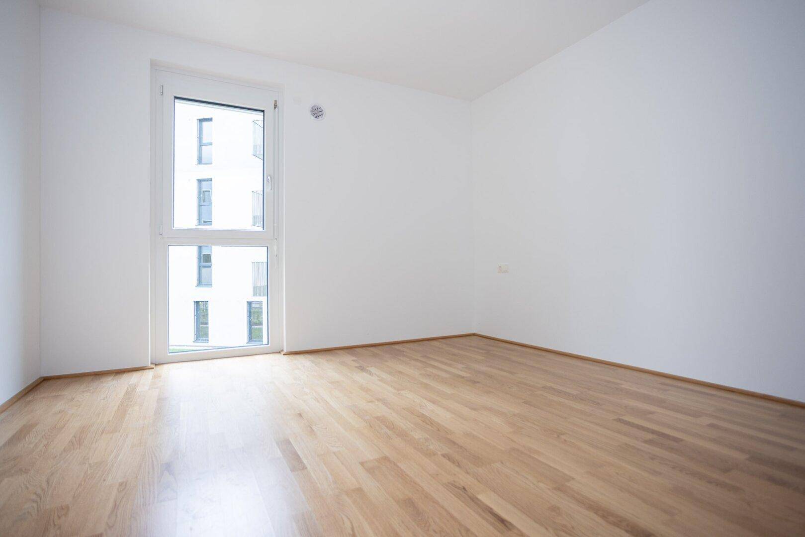 Modernes, helles Zimmer mit Parkettboden und bodentiefem Fenster, das viel Licht spendet.