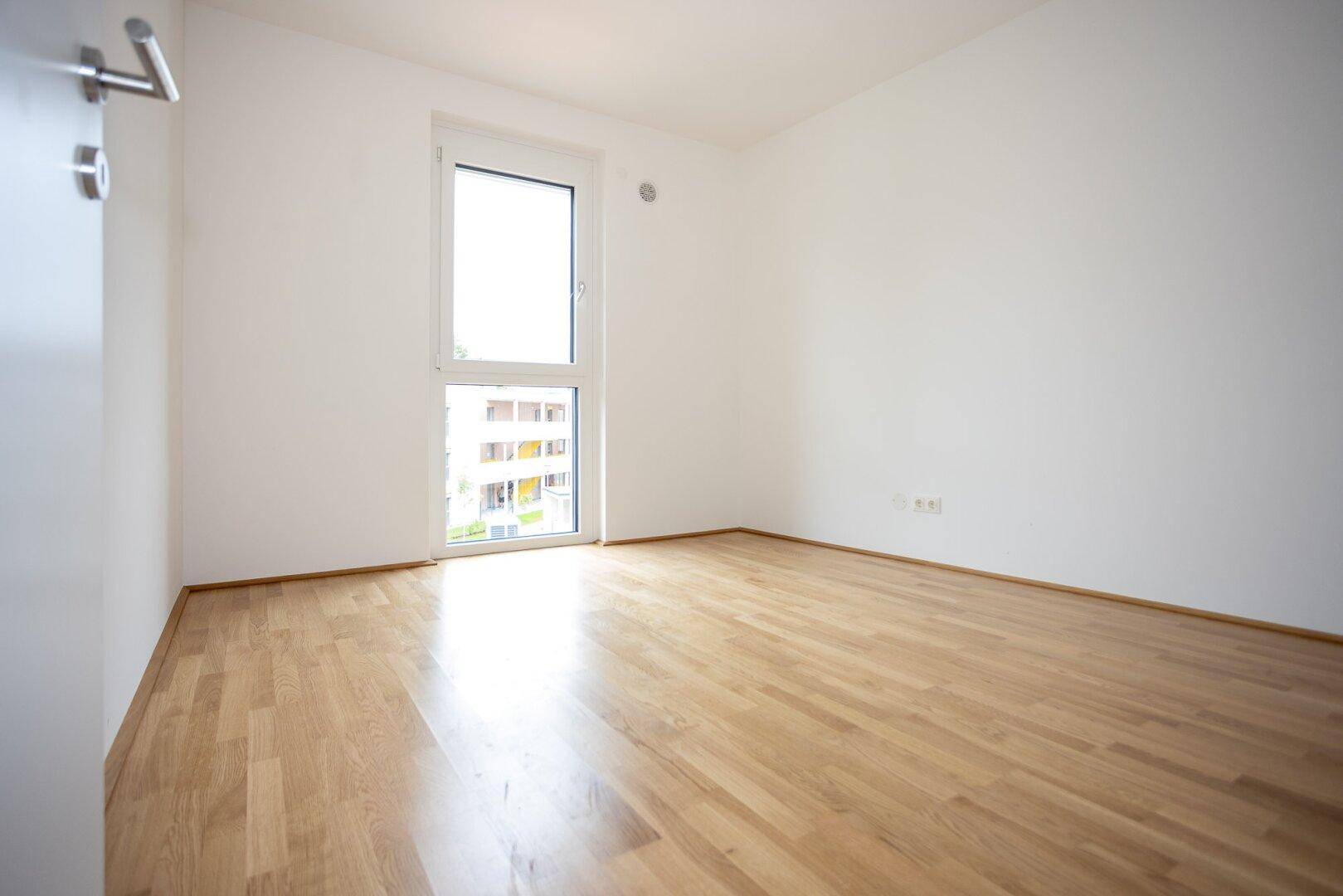 Helles Schlafzimmer mit Parkettboden und großem Fenster, das viel Licht spendet.