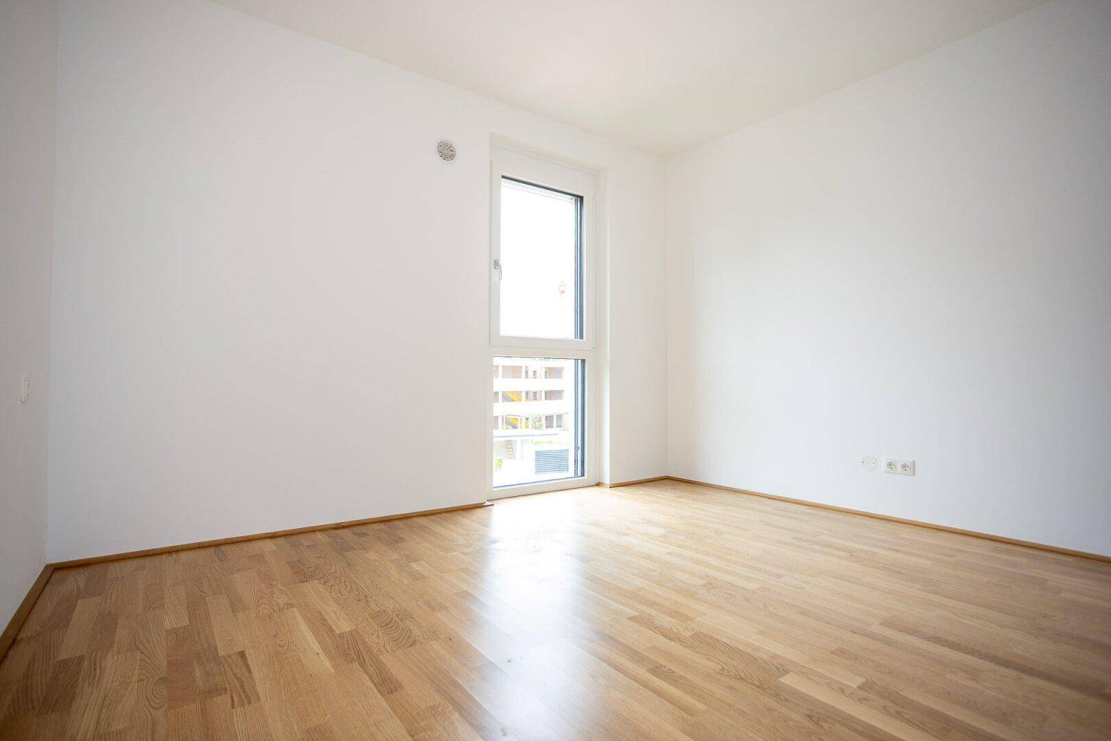 Freundliches Schlafzimmer mit Parkettboden und einem großen Fenster, das eine angenehme Atmosphäre schafft.