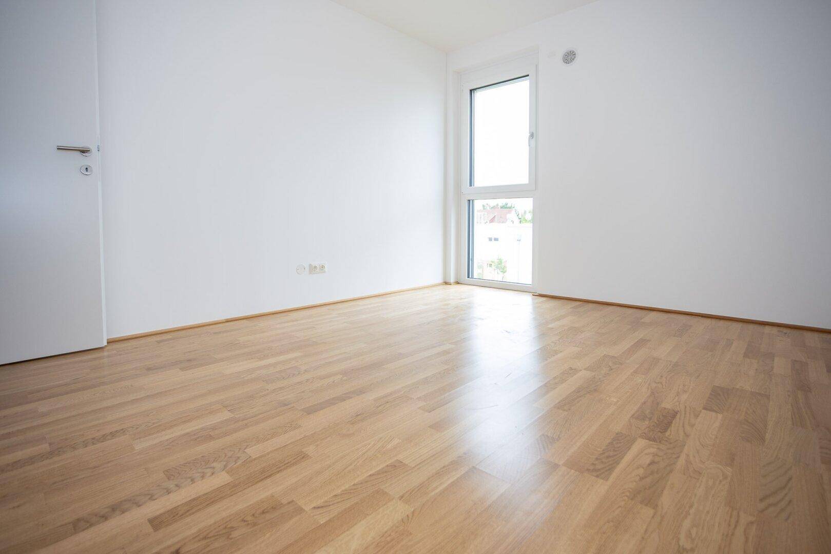 Helles, leeres Schlafzimmer mit Parkettboden und großem Fenster.