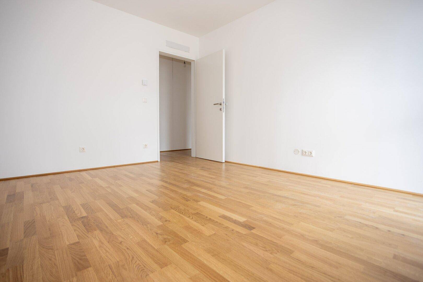 Leeres Schlafzimmer mit Parkettboden und offener Tür zu einem weiteren Raum.