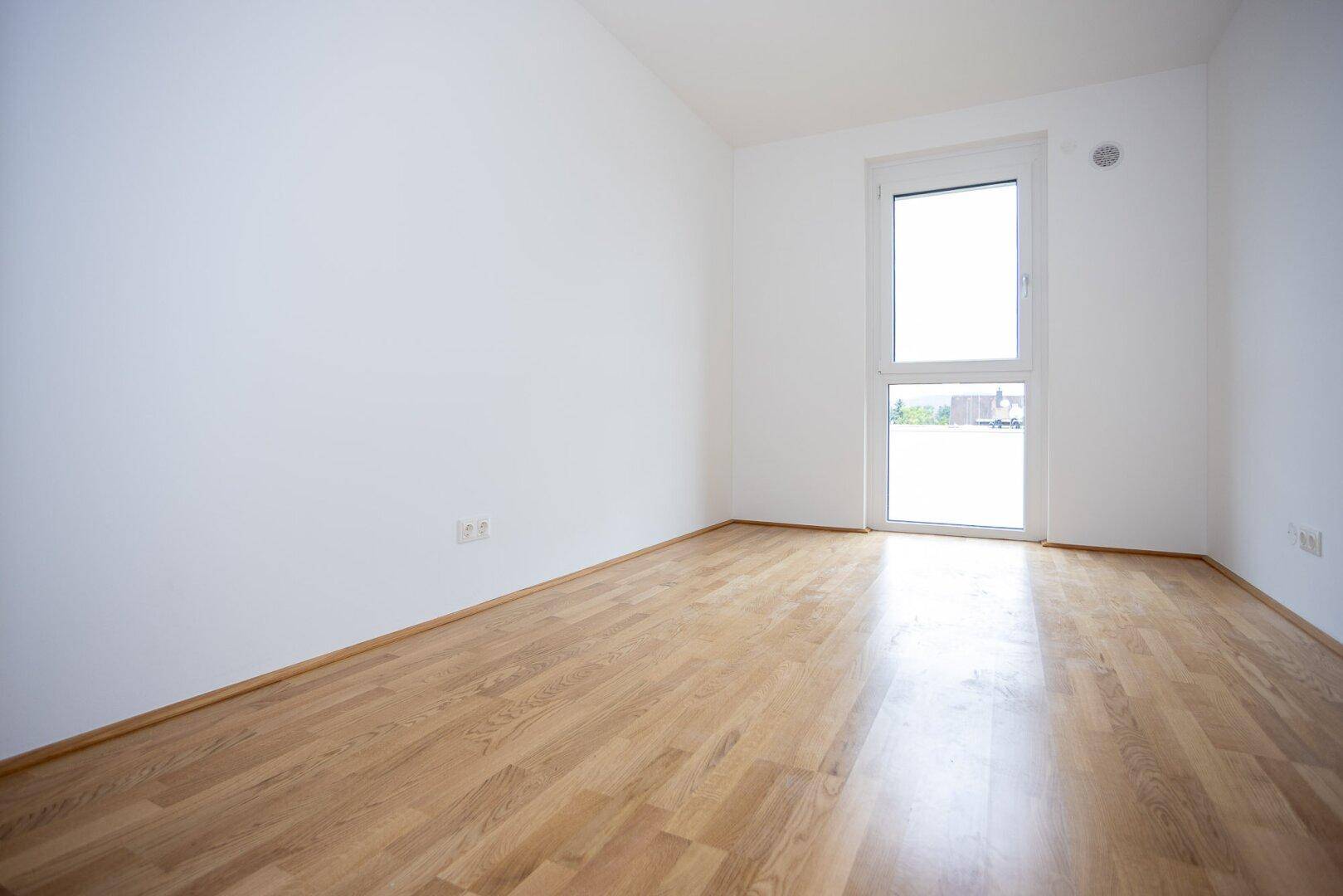 Helles, leeres Schlafzimmer mit Parkettboden und Fenster mit Blick nach draußen.