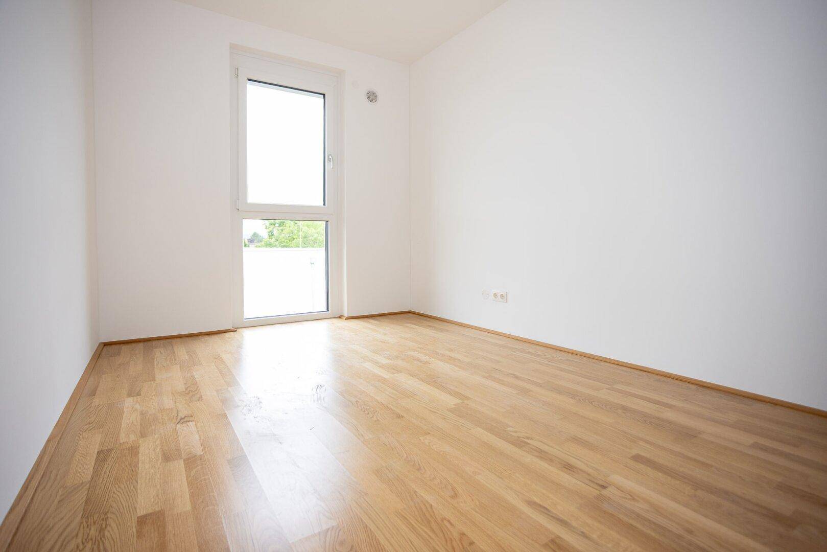 Leeres Schlafzimmer mit Parkettboden und großem Fenster für viel Tageslicht.