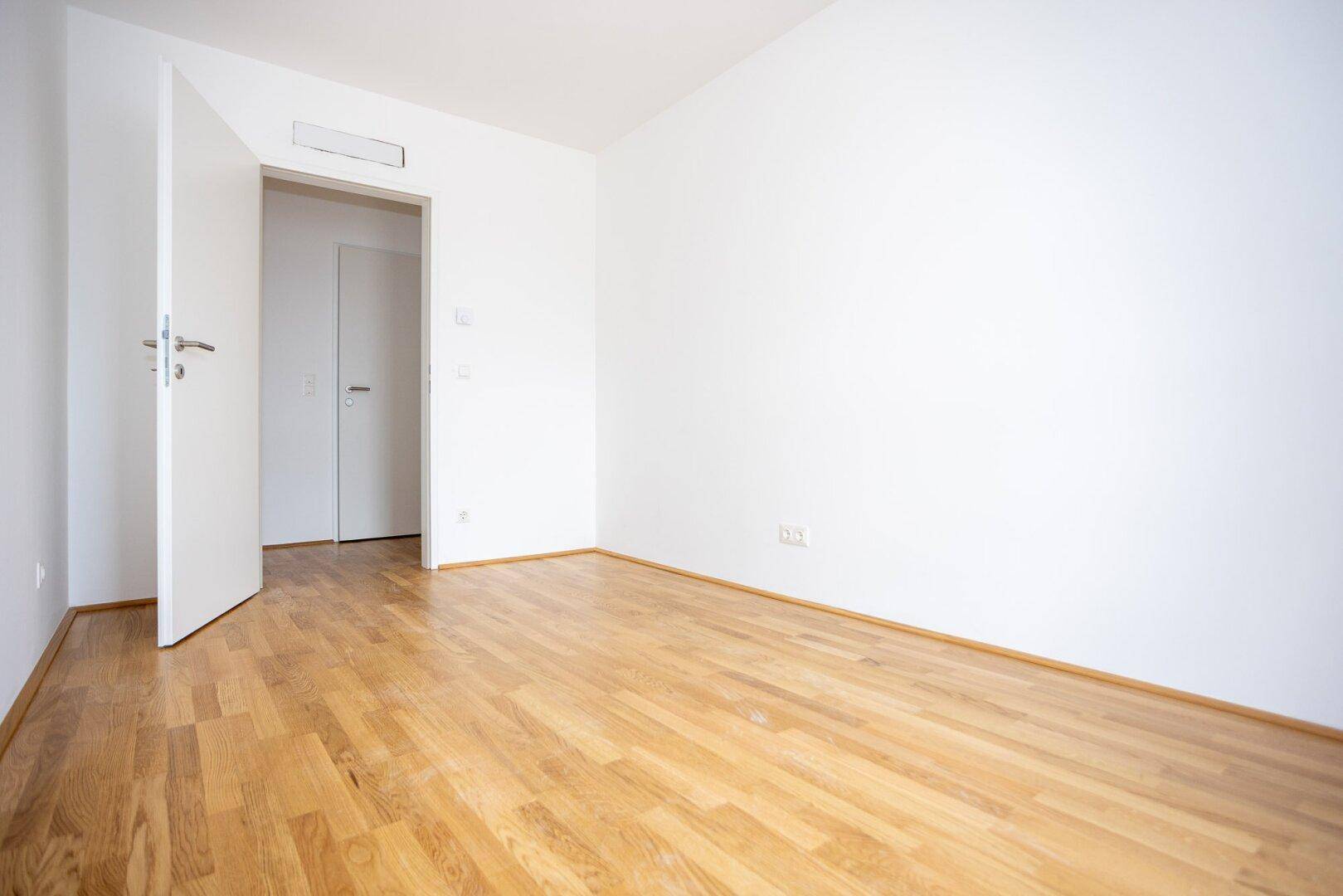 Helles Schlafzimmer mit Parkettboden und offener Tür zu einem angrenzenden Raum.