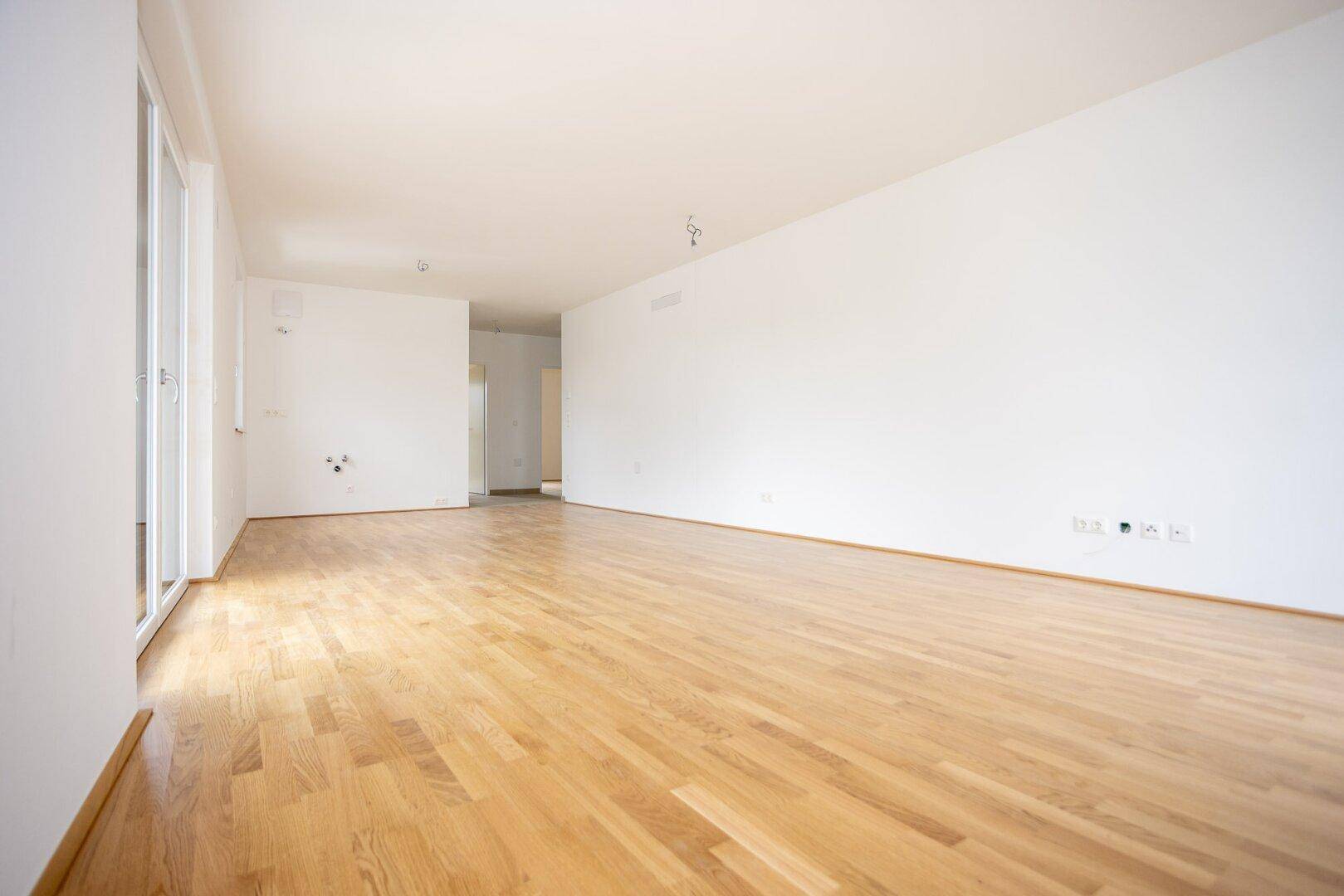 Geräumiges, unmöbliertes Wohnzimmer mit hellem Parkettboden und weiß gestrichenen Wänden.