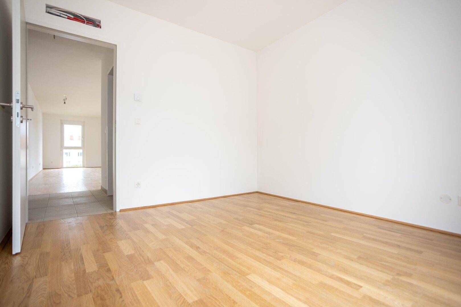Unmöbliertes Zimmer mit Holzboden und Zugang zu einem gefliesten Flur.