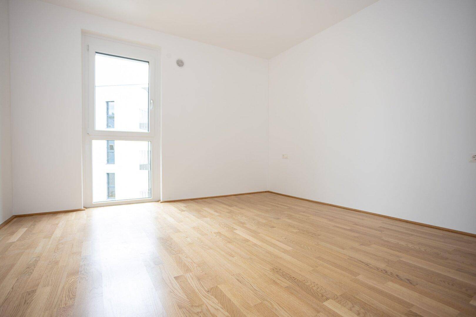 Leeres Zimmer mit Parkettboden und bodentiefem Fenster für viel Tageslicht.