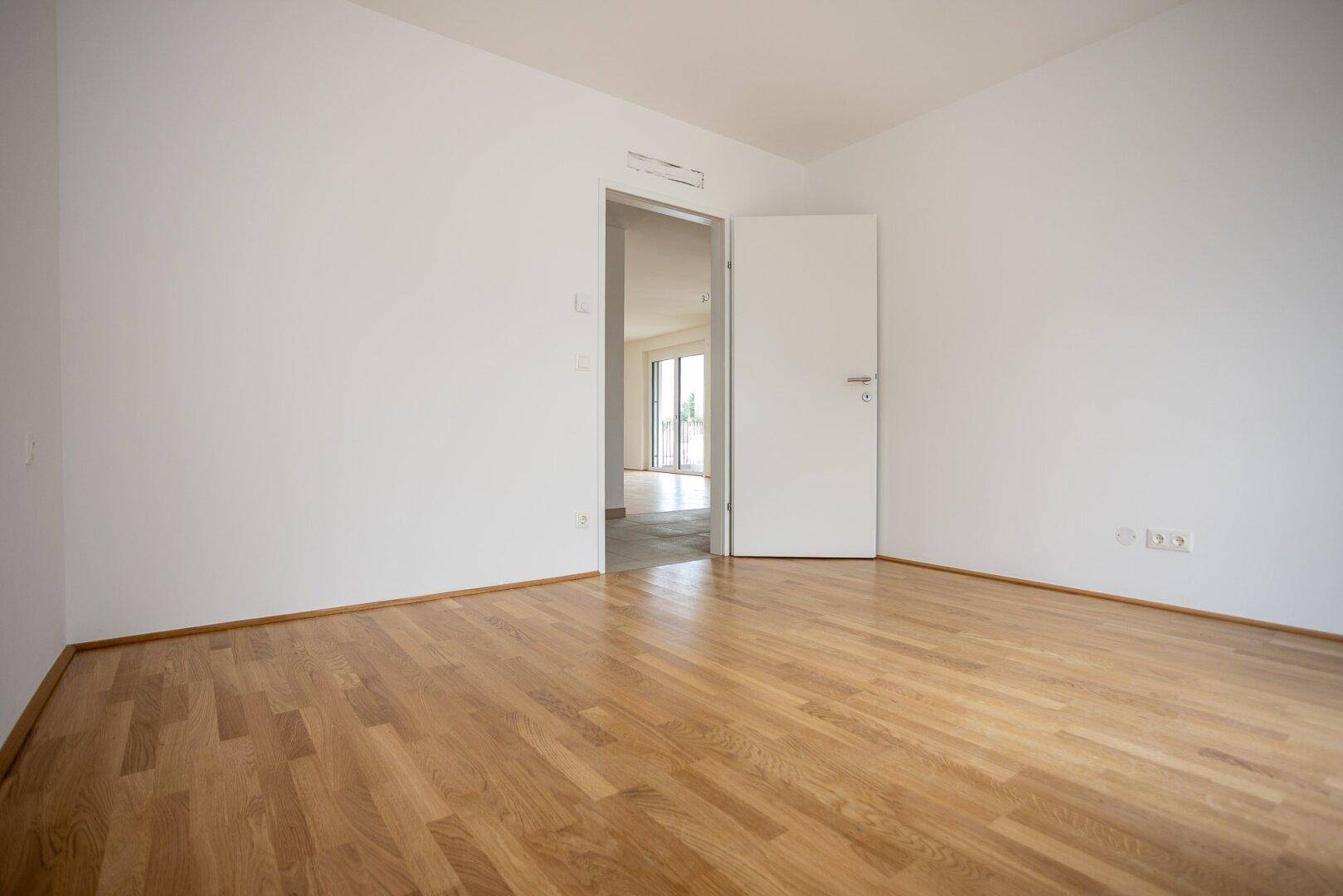 Unmöbliertes Zimmer mit Parkettboden und offener Tür zu einem weiteren Raum.