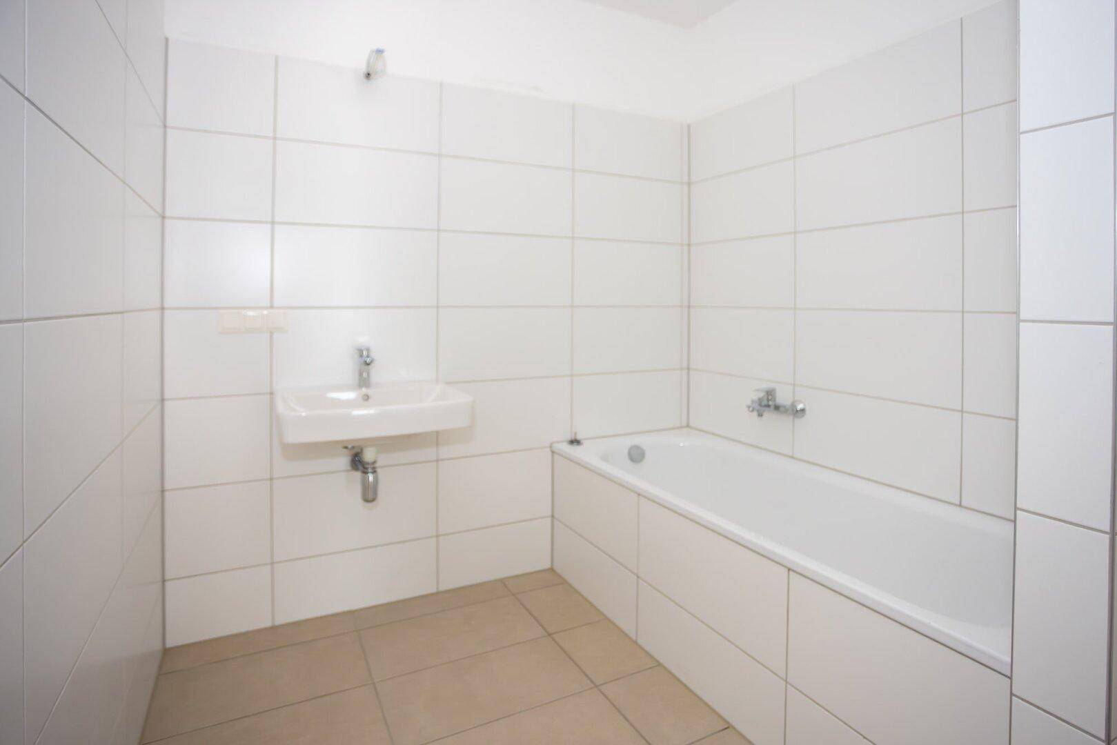 Helles Badezimmer mit weißen Wandfliesen, Badewanne und kleinem Waschbecken.
