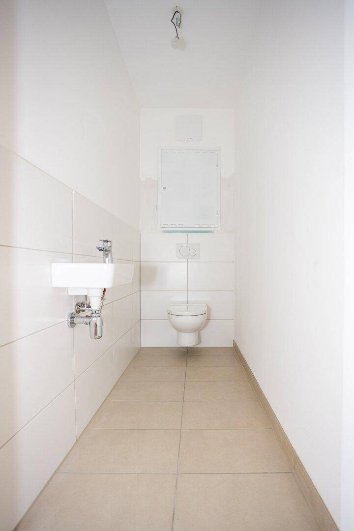 Schmale Gästetoilette mit Wand-WC, kleinem Waschbecken und hellen Fliesen.
