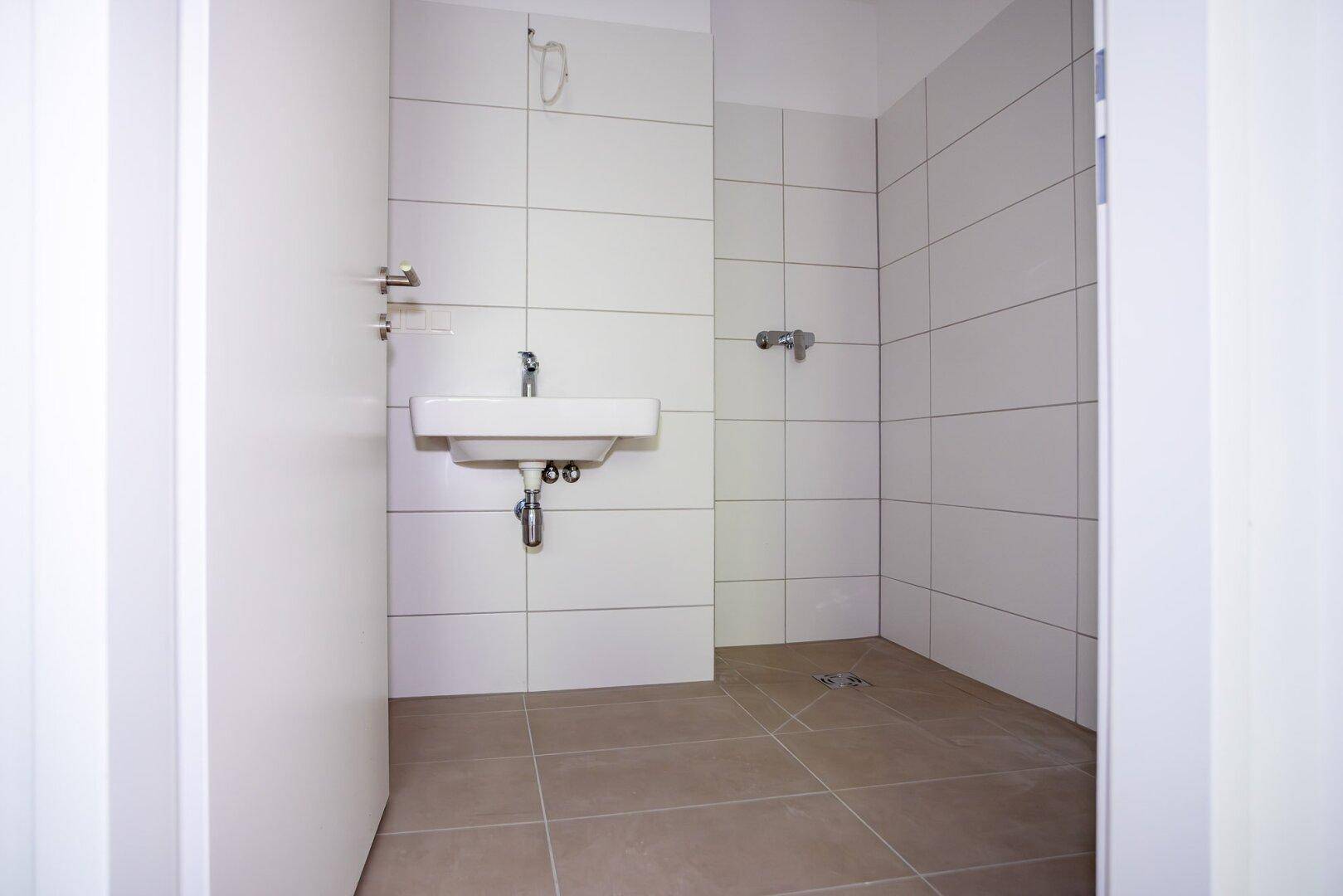 Modern gefliestes Badezimmer mit Waschbecken und vorbereitetem Duschbereich in neutralen Farben.