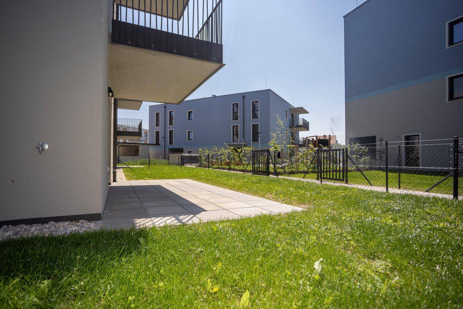 Neubauwohnungen mit privaten Terrassen und umzäunten Gartenbereichen an einem sonnigen Tag.