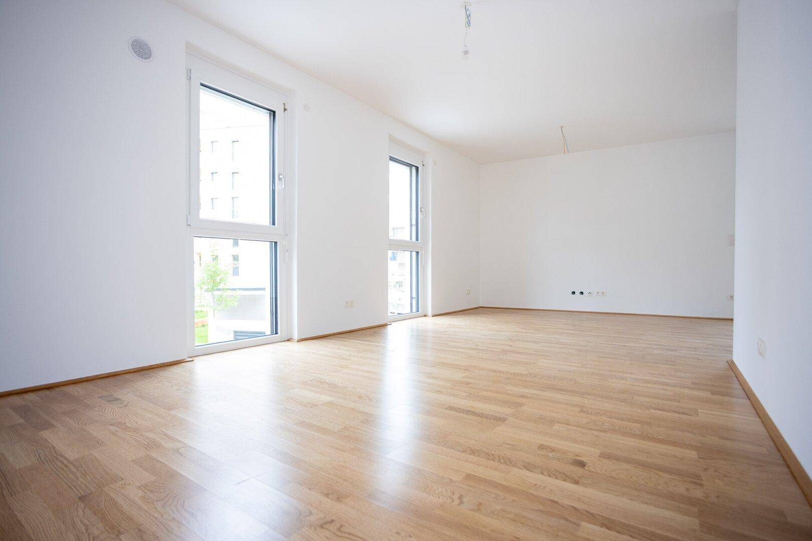 Lichtdurchfluteter Raum mit großen Fenstern und hellem Parkettboden, ideal für ein Wohnzimmer.
