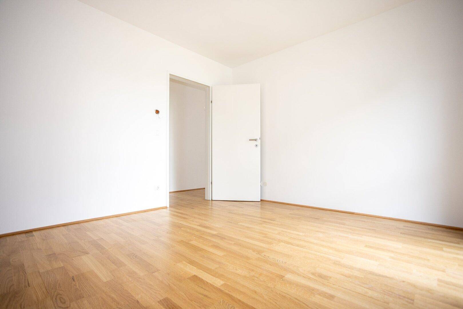 Geräumiges Zimmer mit hellem Parkettboden und offener Tür zu einem weiteren Raum.