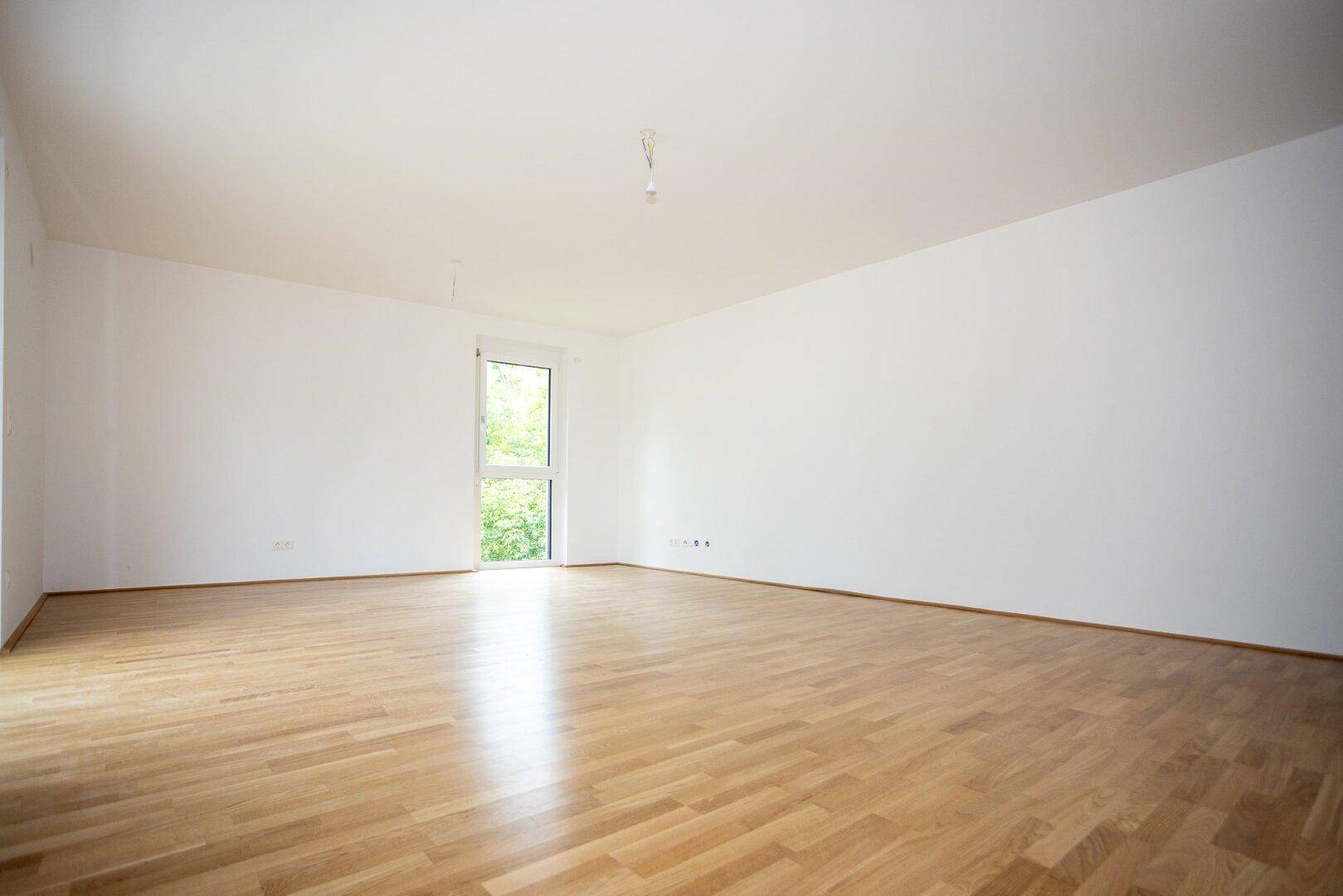Helles Zimmer mit Holzboden und einem großen Fenster, das einen Blick ins Grüne bietet.