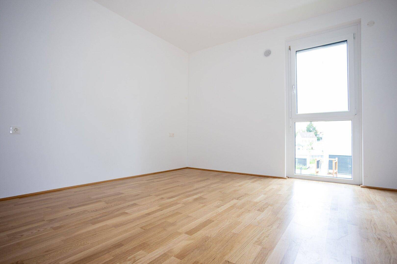 Leeres Zimmer mit hellem Holzboden und einem Fenster, das viel Tageslicht hereinlässt.