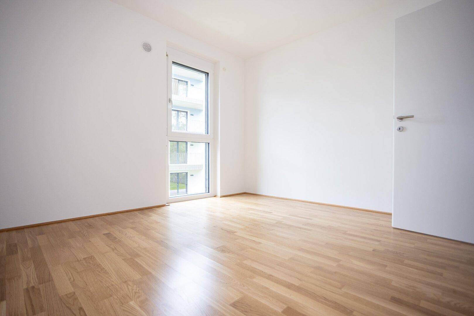 Helles Zimmer mit Holzboden und einem Fenster, das den Blick auf ein Nachbargebäude freigibt.