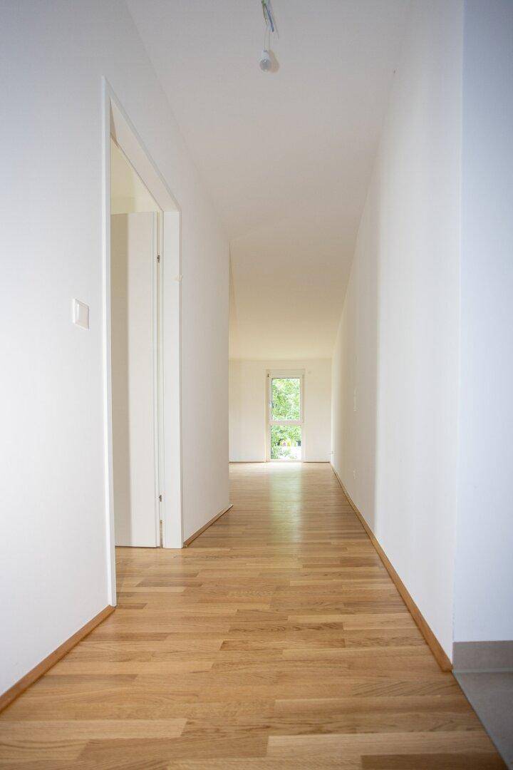 Langer, heller Flur mit Holzboden und weißen Wänden, der zu einem Fenster am Ende führt.