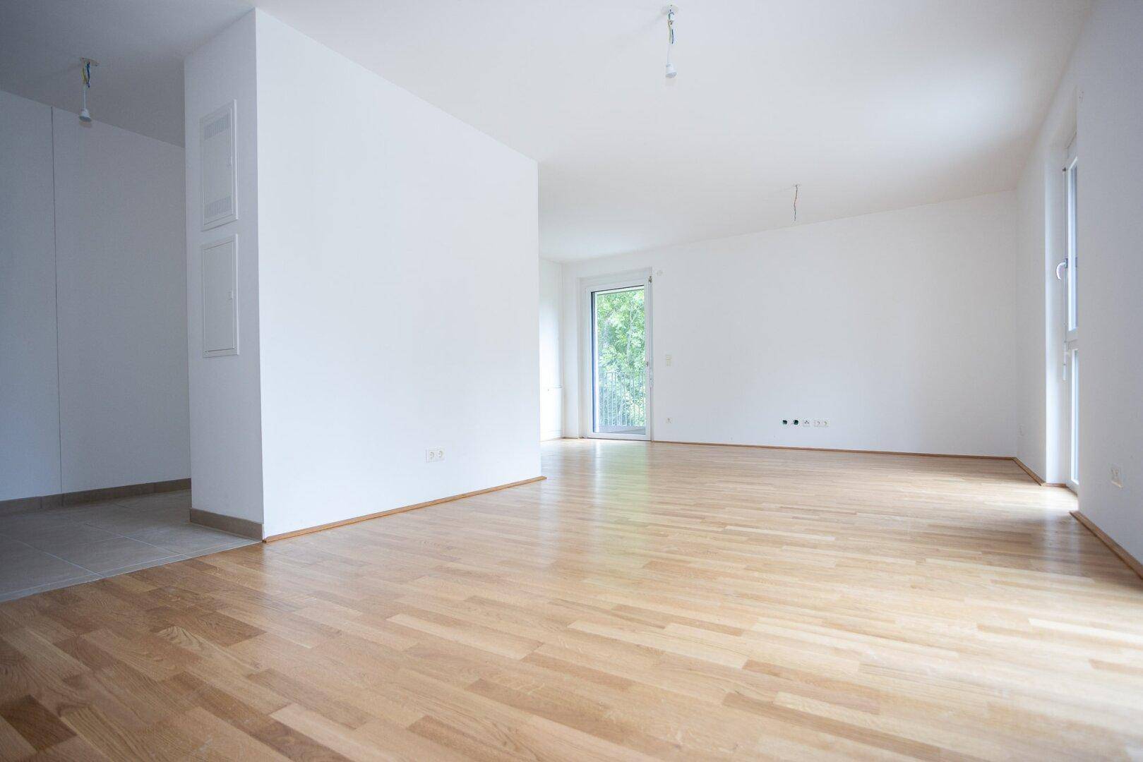 Leeres Zimmer mit hellem Parkettboden und Zugang zu einem Außenbereich durch eine Glastür.
