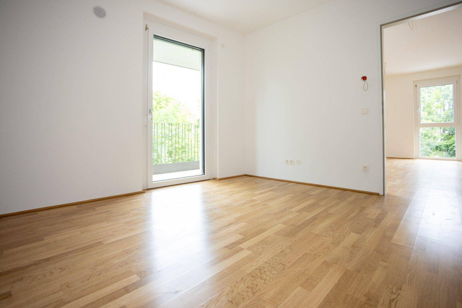 Lichtdurchflutetes Zimmer mit Parkettboden und Balkonzugang, verbunden mit einem weiteren Raum.