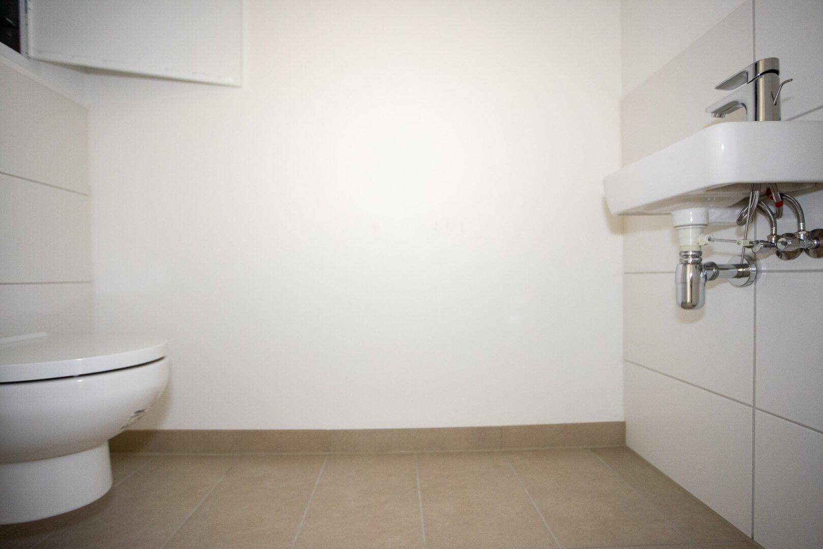 Separates WC mit weißer Toilette, kleinem Waschbecken und hellen Wandfliesen.