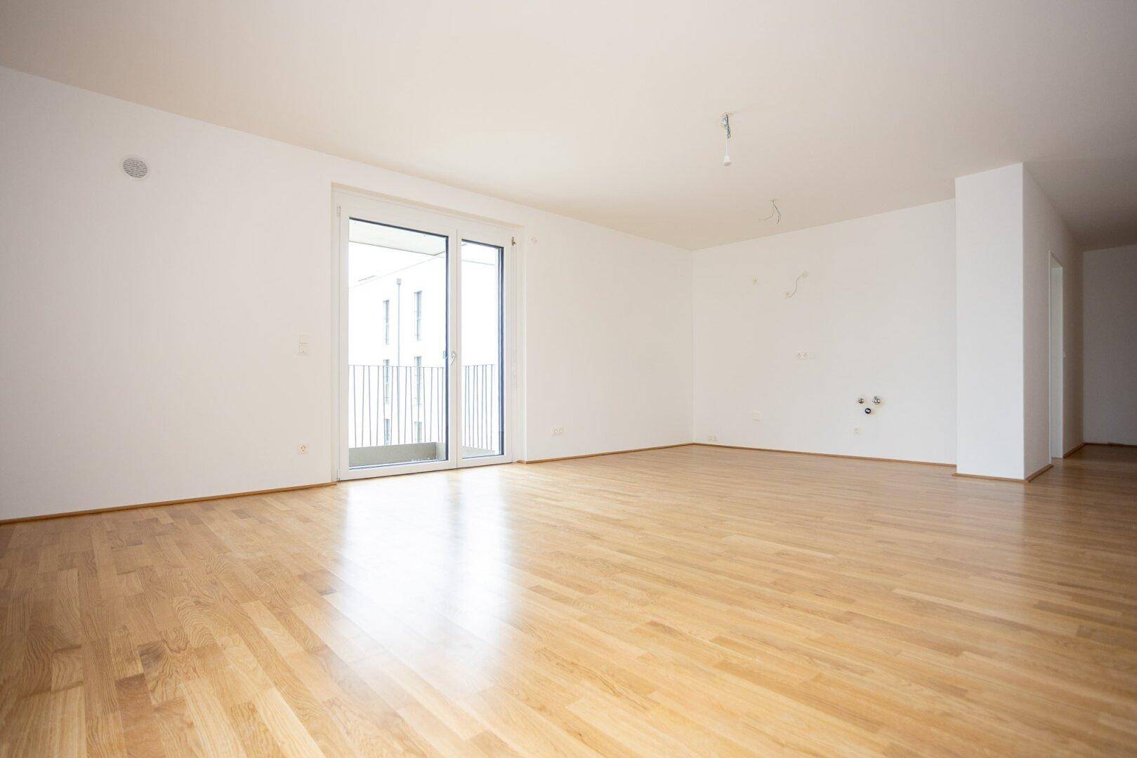 Heller, leerer Raum mit Parkettboden und Schiebetür, die zu einem Balkon führt.